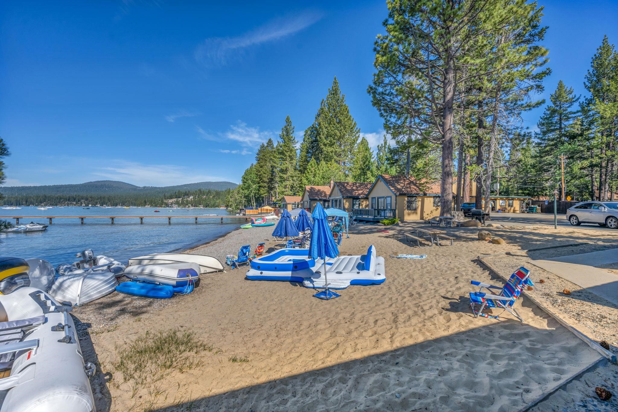 Sandy lakeside beach with inflatable boats and a dock beside pine-shaded cabins.