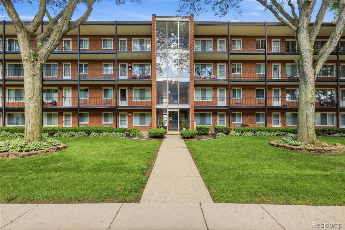 Red-brick apartment building with balconies, a glass entrance, and a walkway across the lawn.