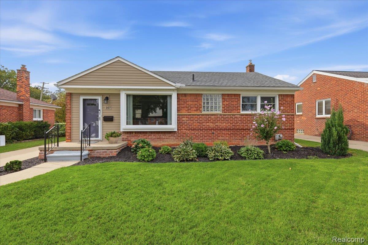 Single-story brick house with tan entry siding, landscaped flower beds, and a broad green lawn.