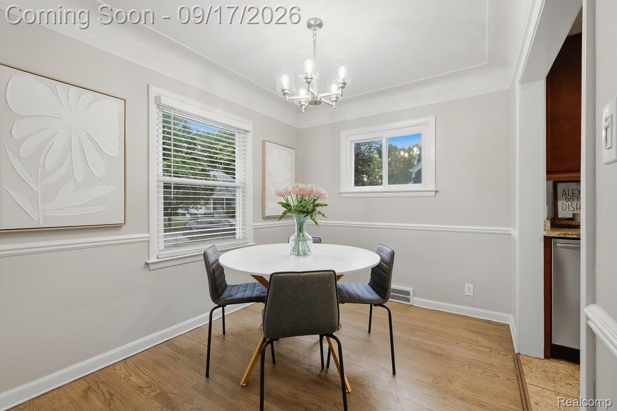 Dining room with a round white table, four chairs, and a vase of pink flowers by the windows.