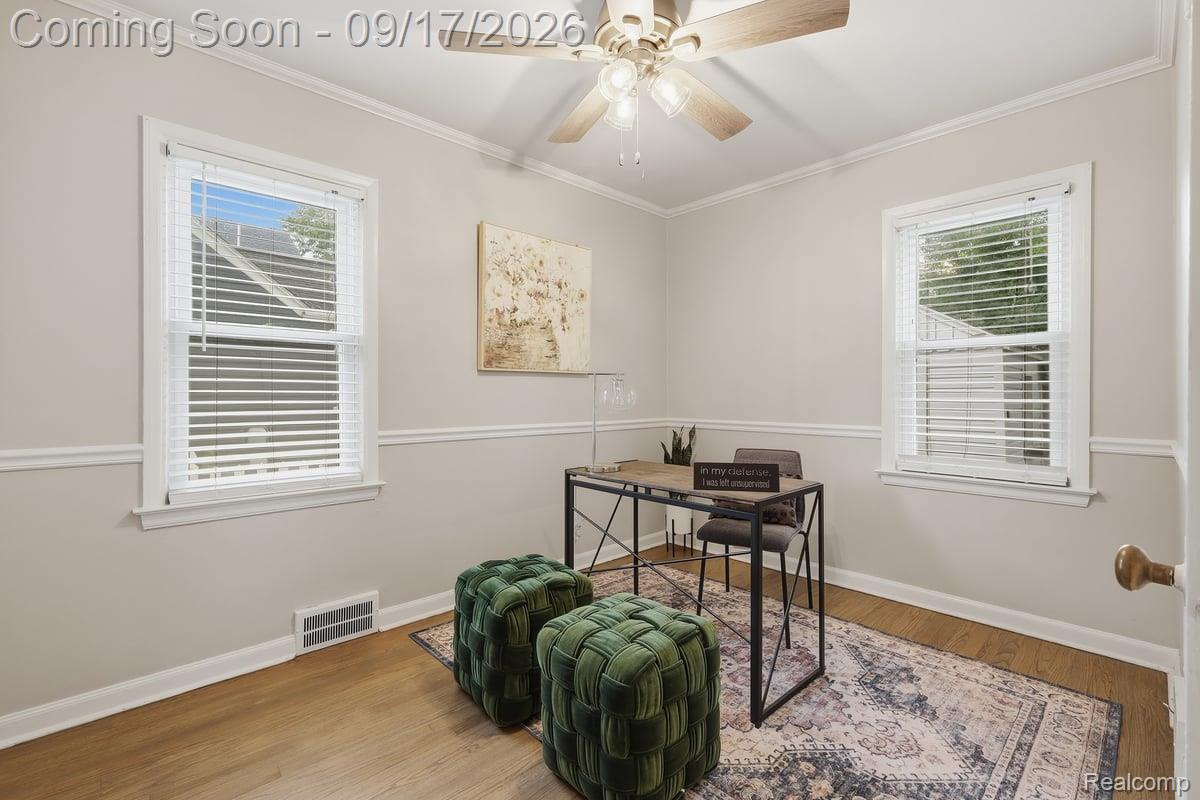 Staged home office with two bright windows, a desk, patterned rug, and dark green tufted ottomans.