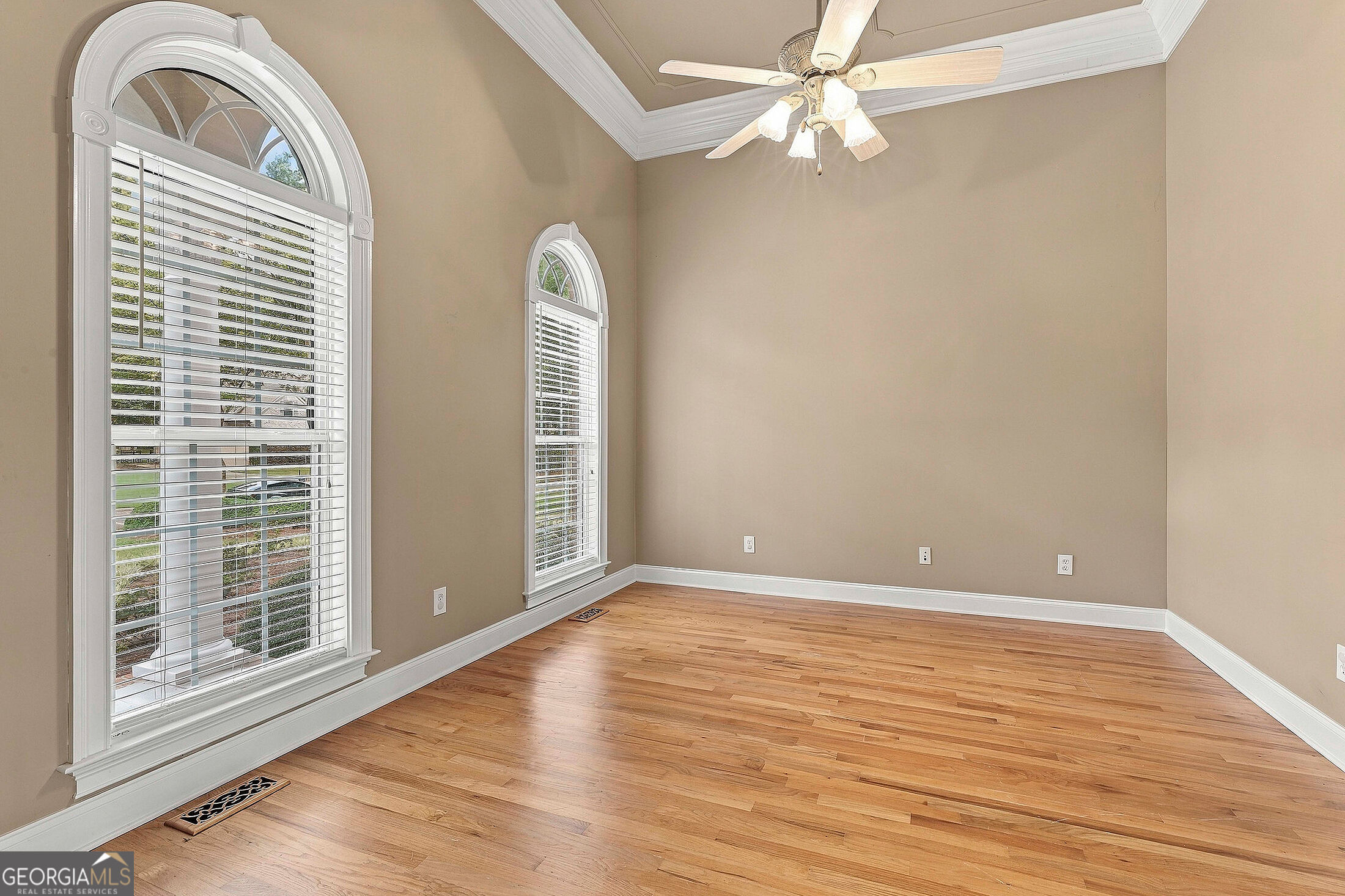 Empty room with hardwood floors, a ceiling fan, and two arched windows with blinds.