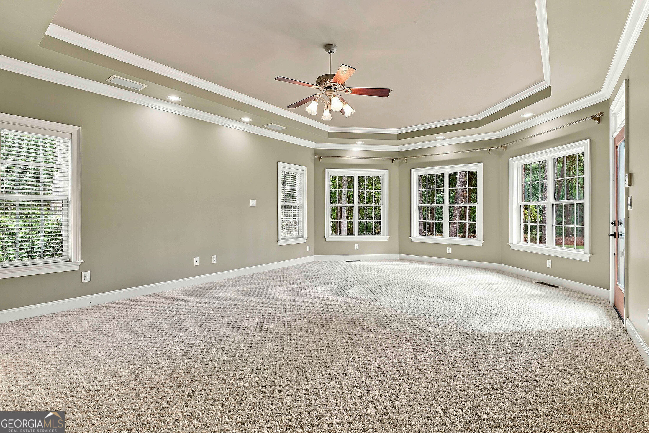 Empty living room with a tray ceiling, ceiling fan, and multiple windows overlooking trees.