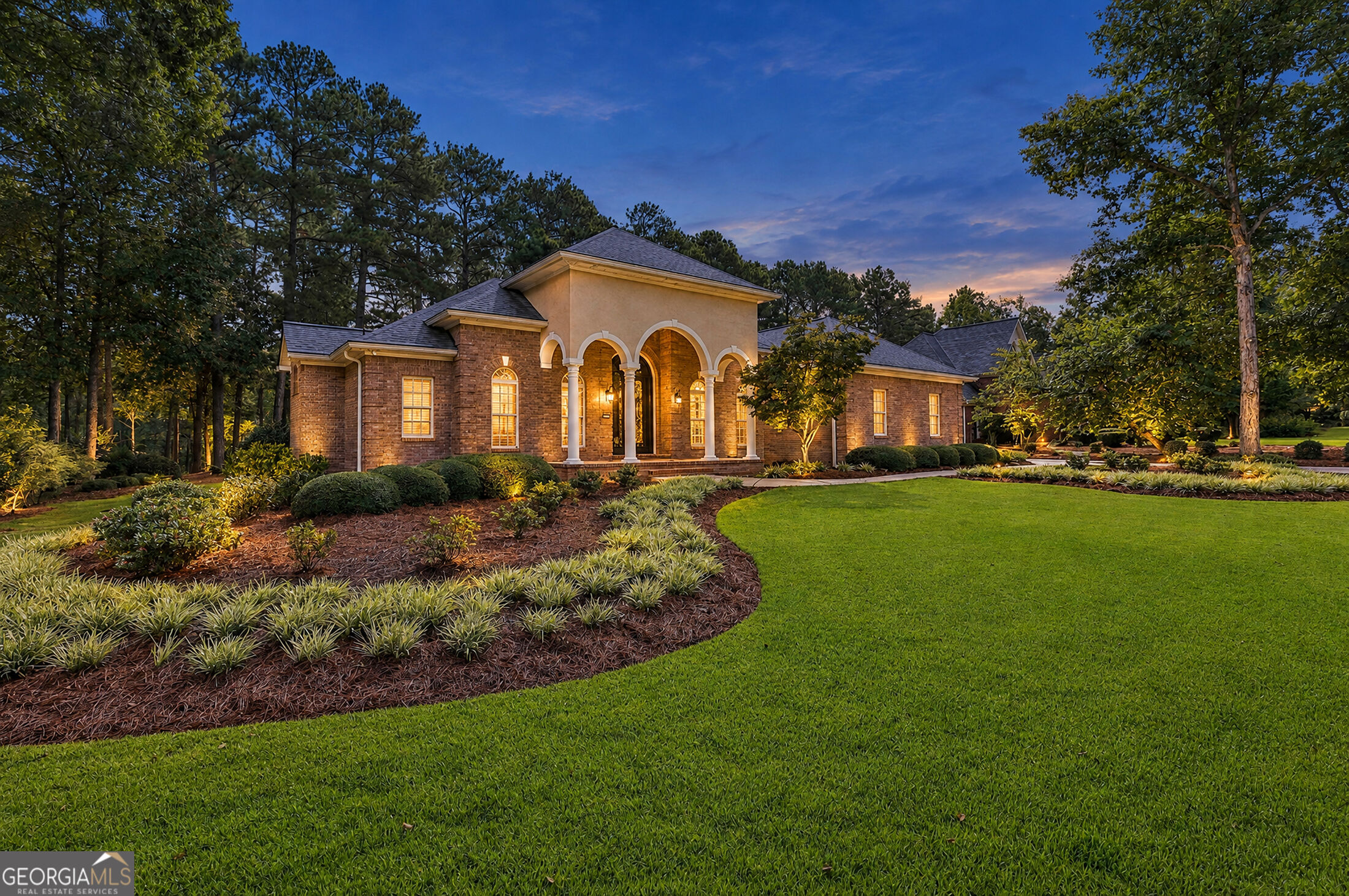 Brick-and-stucco home with arched entry, illuminated landscaping, and broad front lawn at dusk.