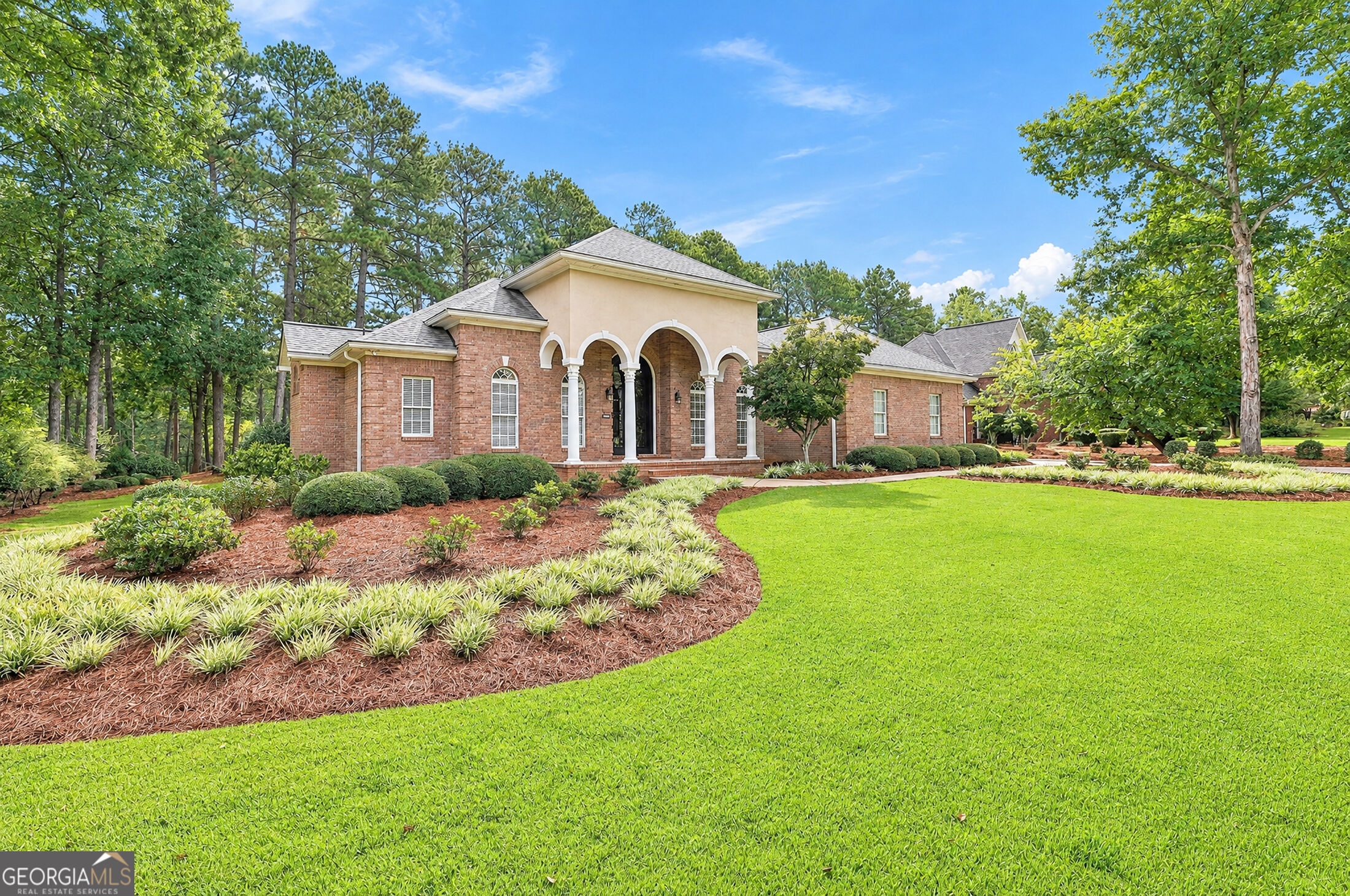 Brick home with arched white-column entry, landscaped beds, and a broad green lawn.