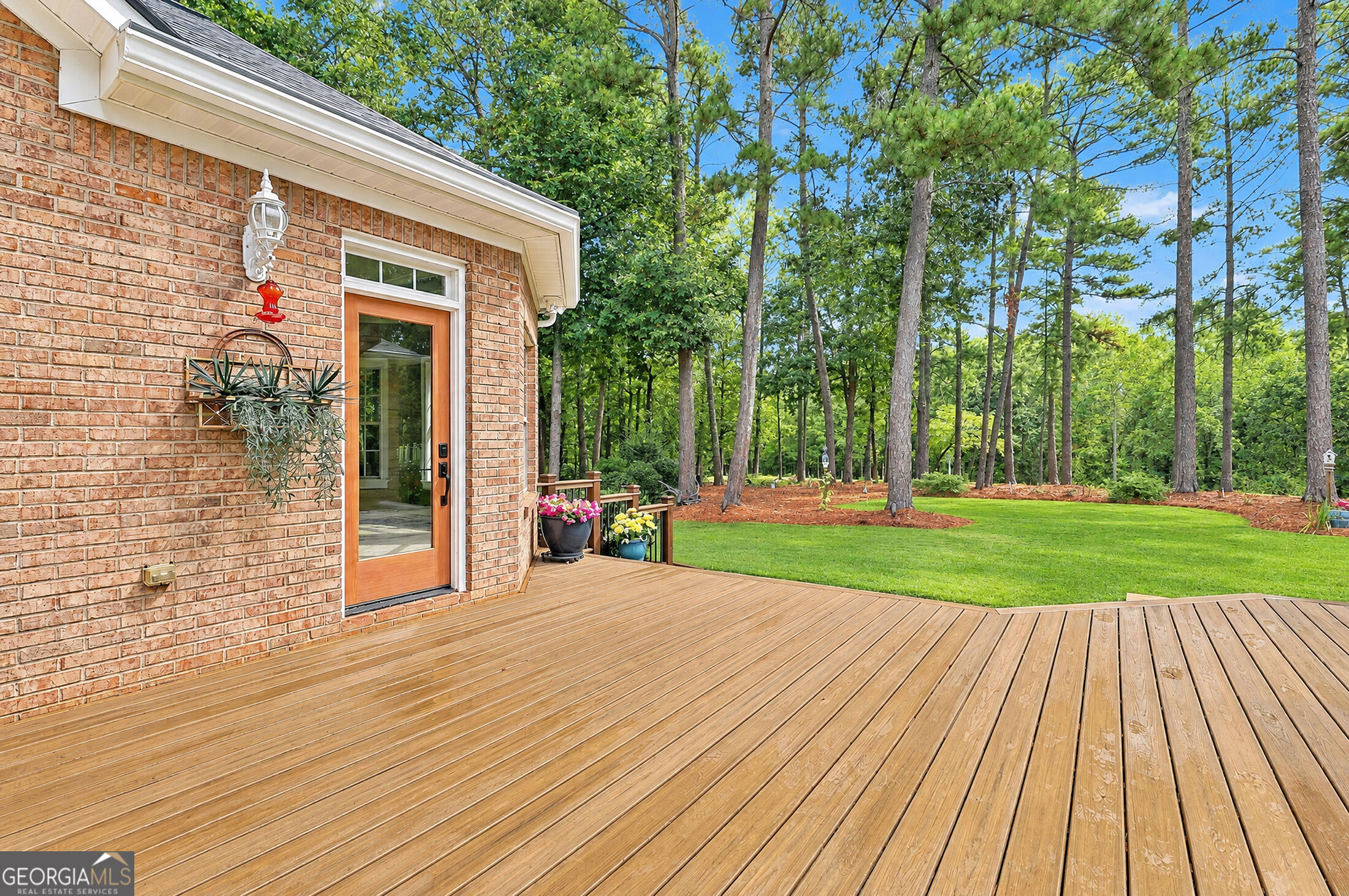 Wooden deck beside a brick home, opening onto a tree-lined lawn.