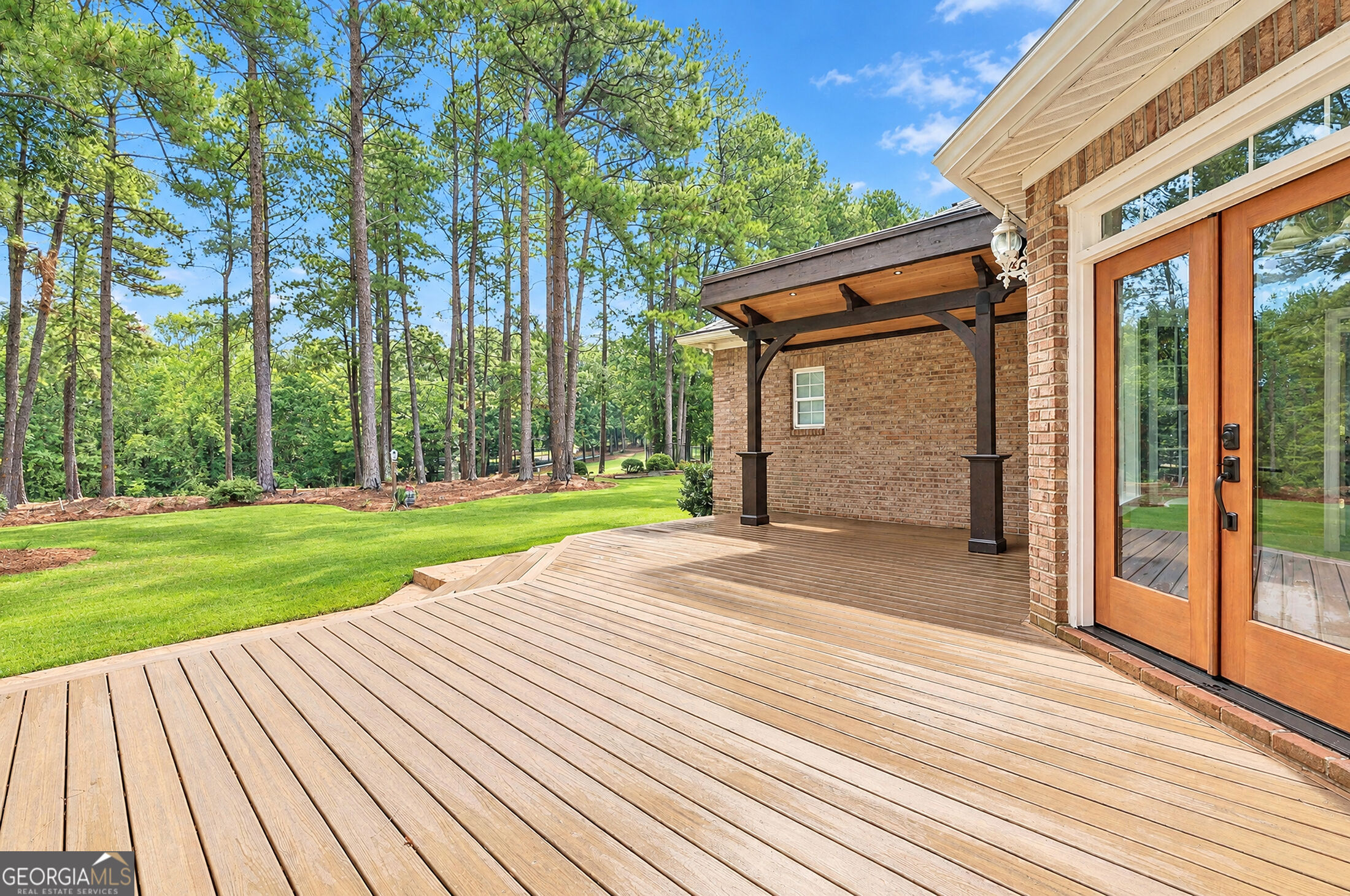 Wooden deck beside a brick home, with a covered patio overlooking a lawn and pine trees.