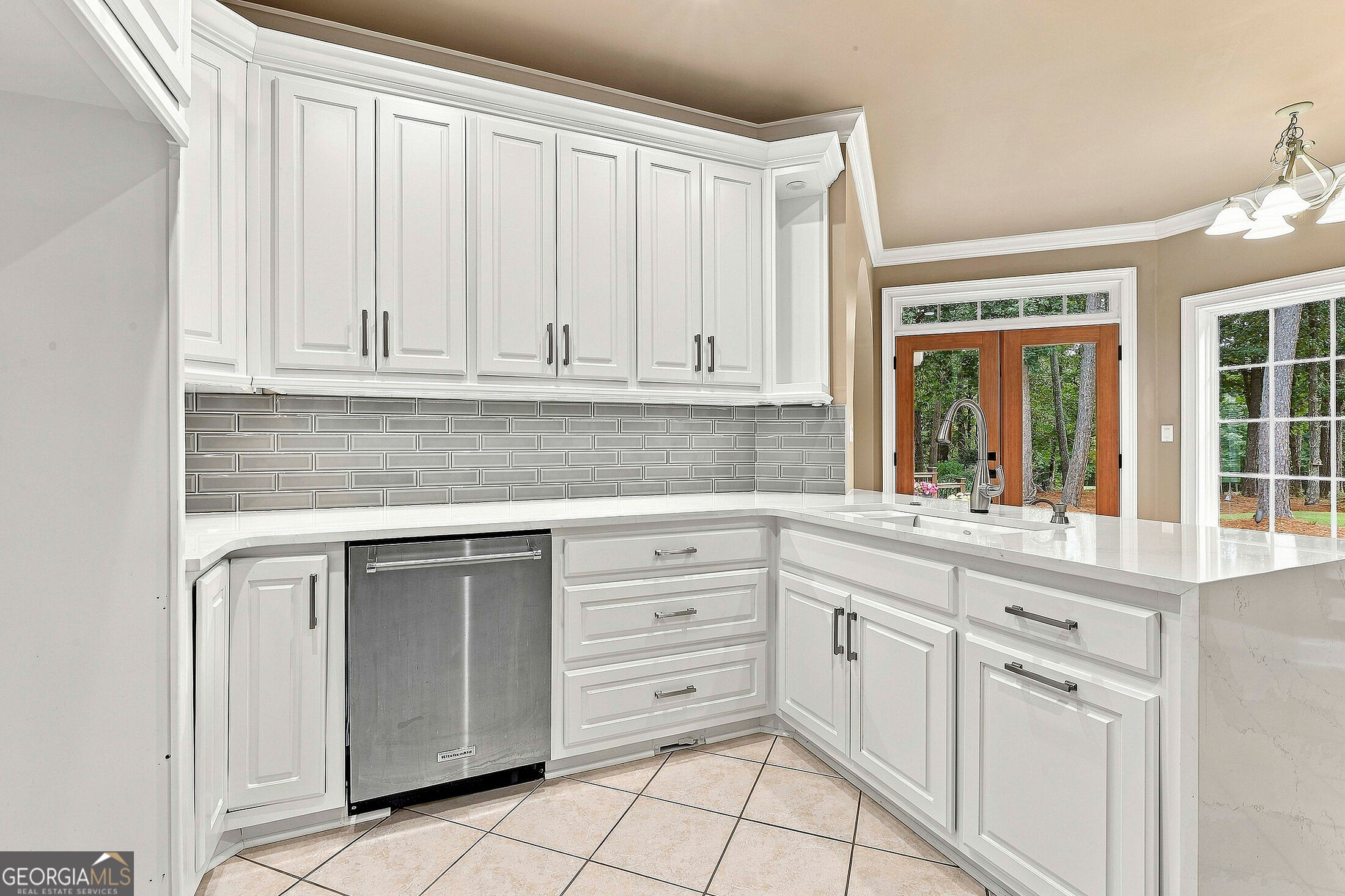 Kitchen with white cabinets, gray tile backsplash, stainless dishwasher, and windows overlooking trees.