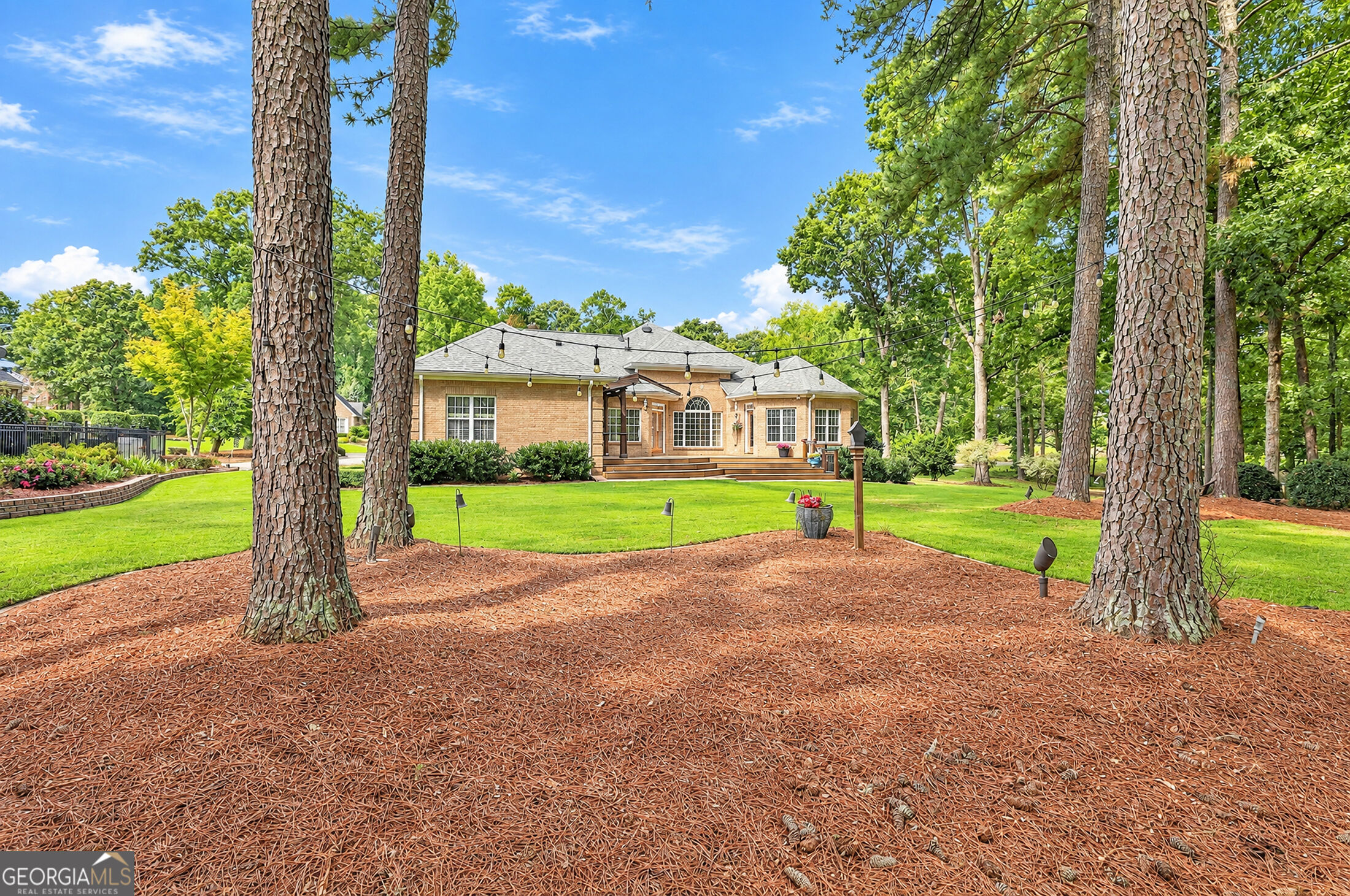Brick home’s rear exterior beyond a broad lawn, framed by mature pines and overhead string lights.