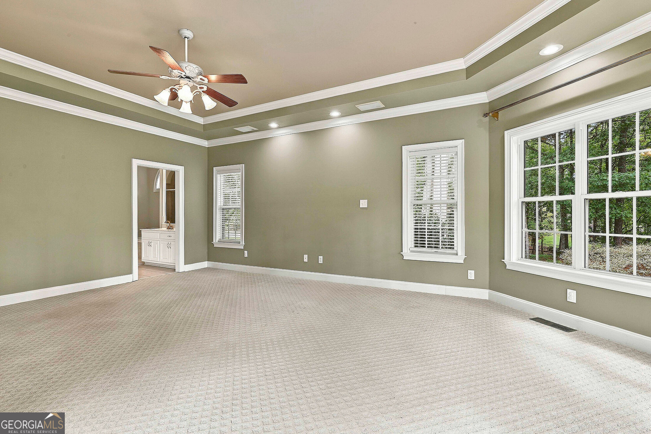 Empty bedroom with beige carpet, tray ceiling, ceiling fan, and windows overlooking trees.