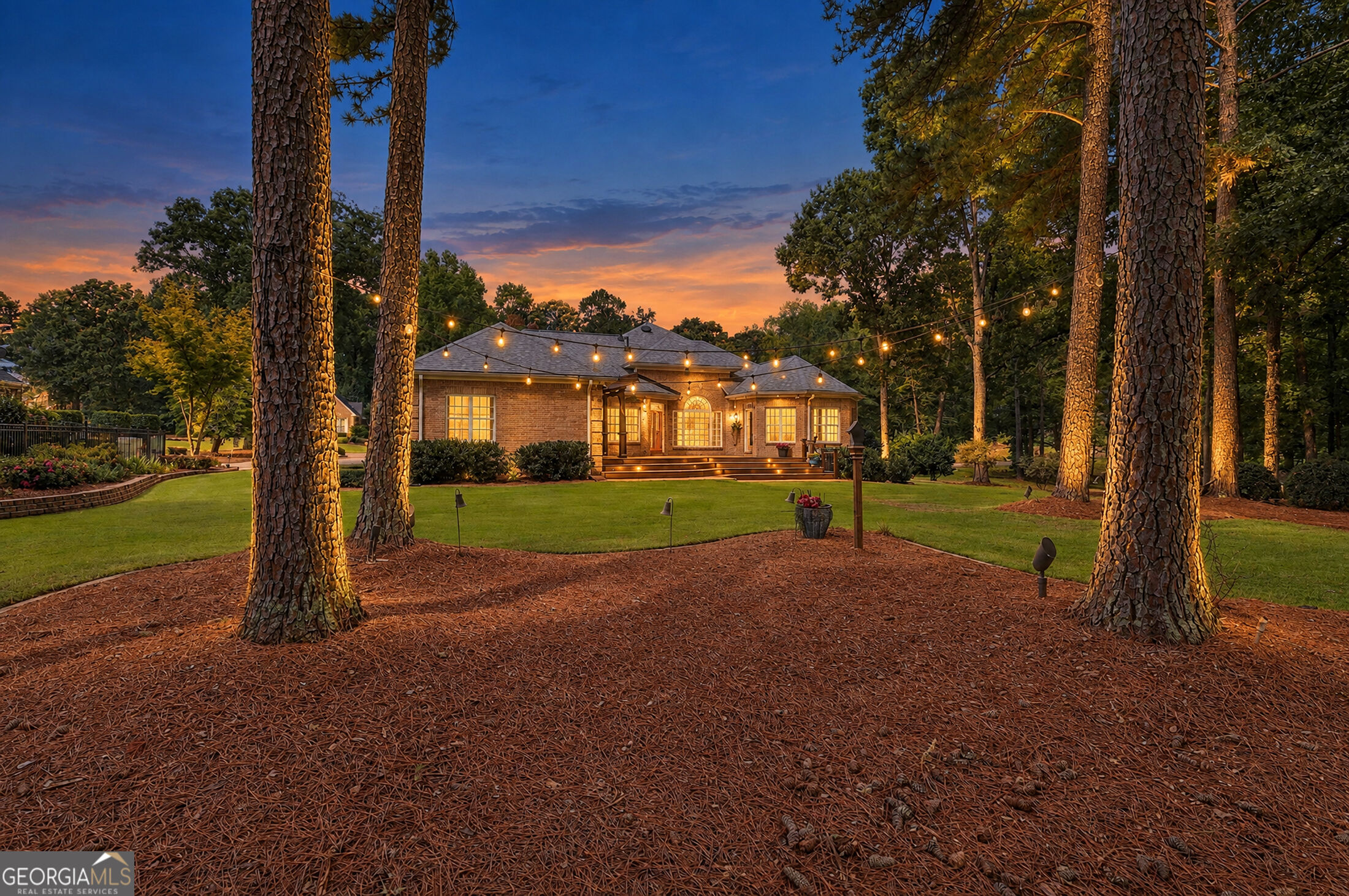 Brick home framed by tall pines, with glowing windows and string lights across the backyard at dusk.
