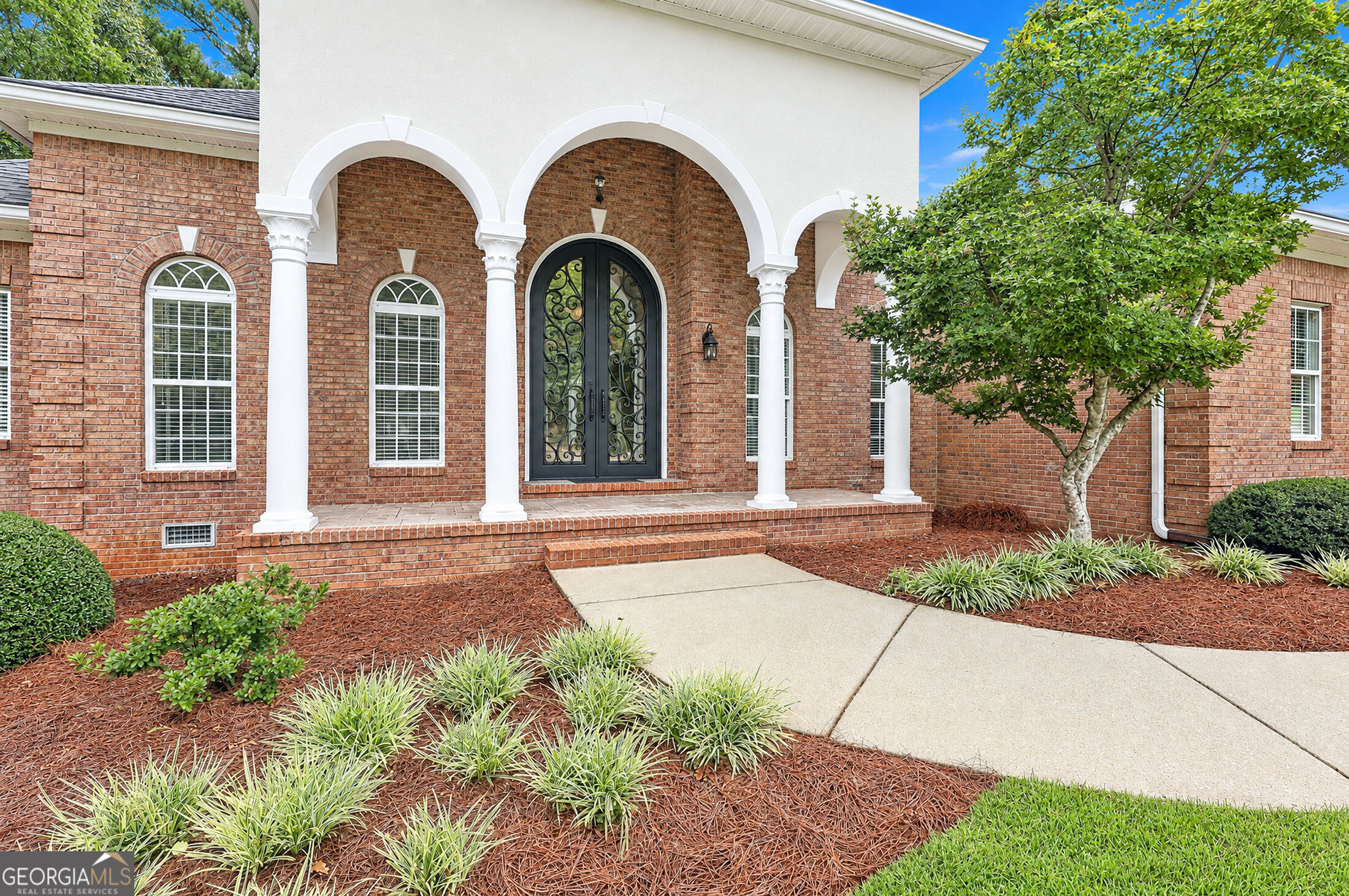 Brick home entrance with a white arched portico, ornate black double doors, and landscaped walkway.