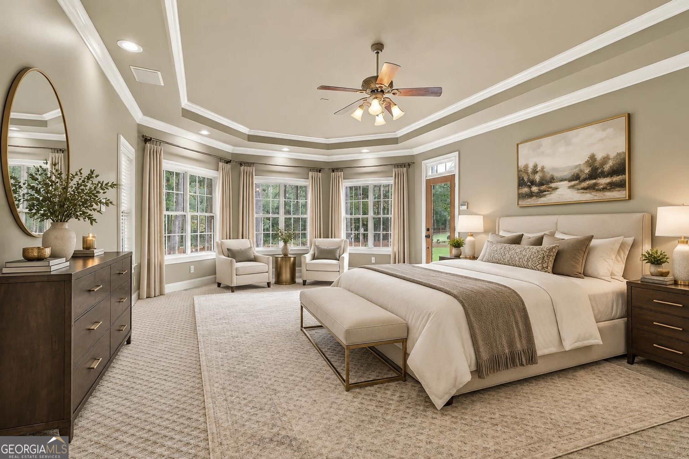 Furnished bedroom with a large bed, tray ceiling, and a bright window-side sitting area.