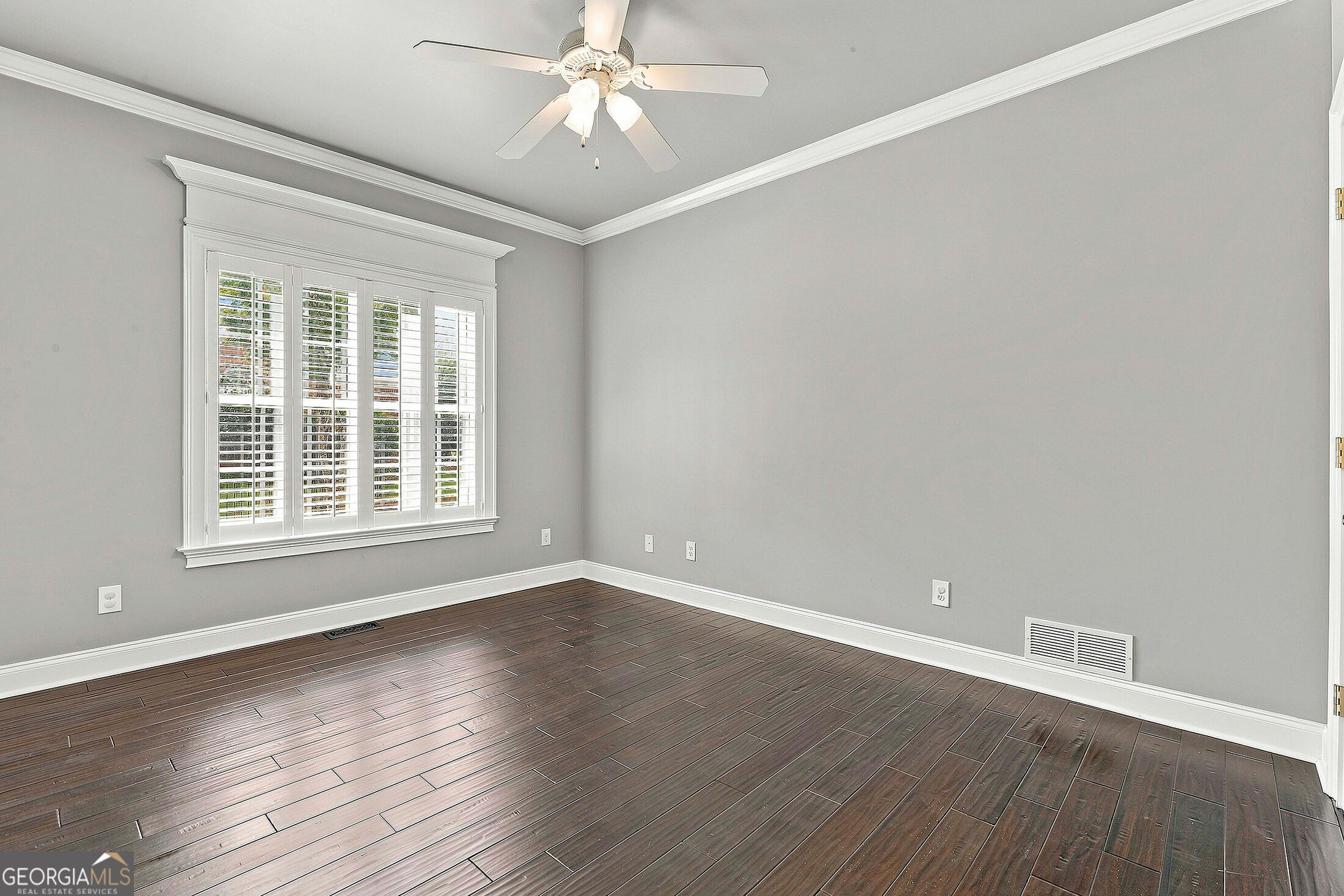 Empty room with dark plank flooring, gray walls, white plantation shutters, and a ceiling fan.
