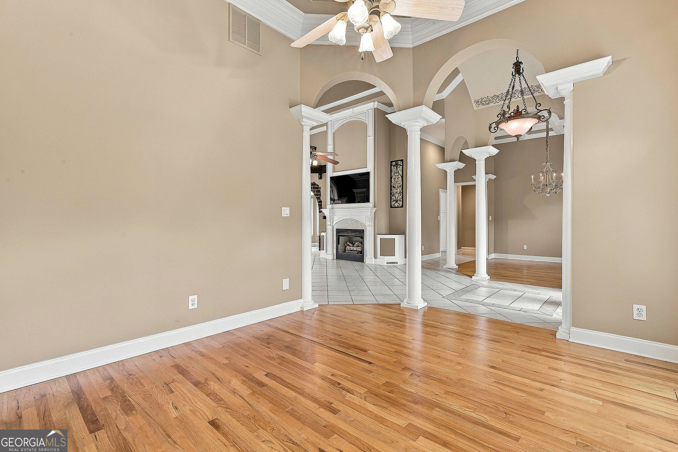 Empty hardwood-floored room with white arches opening to a tiled living area and fireplace.