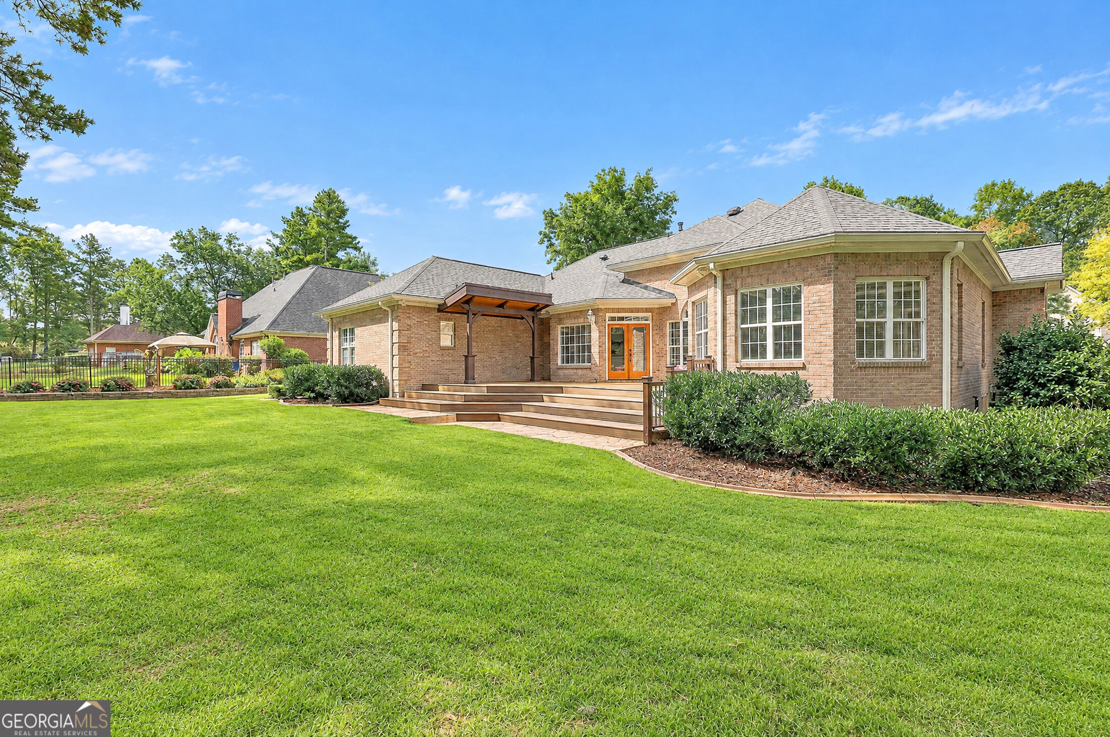 Brick house rear exterior with a covered wooden deck, landscaped shrubs, and a broad lawn.