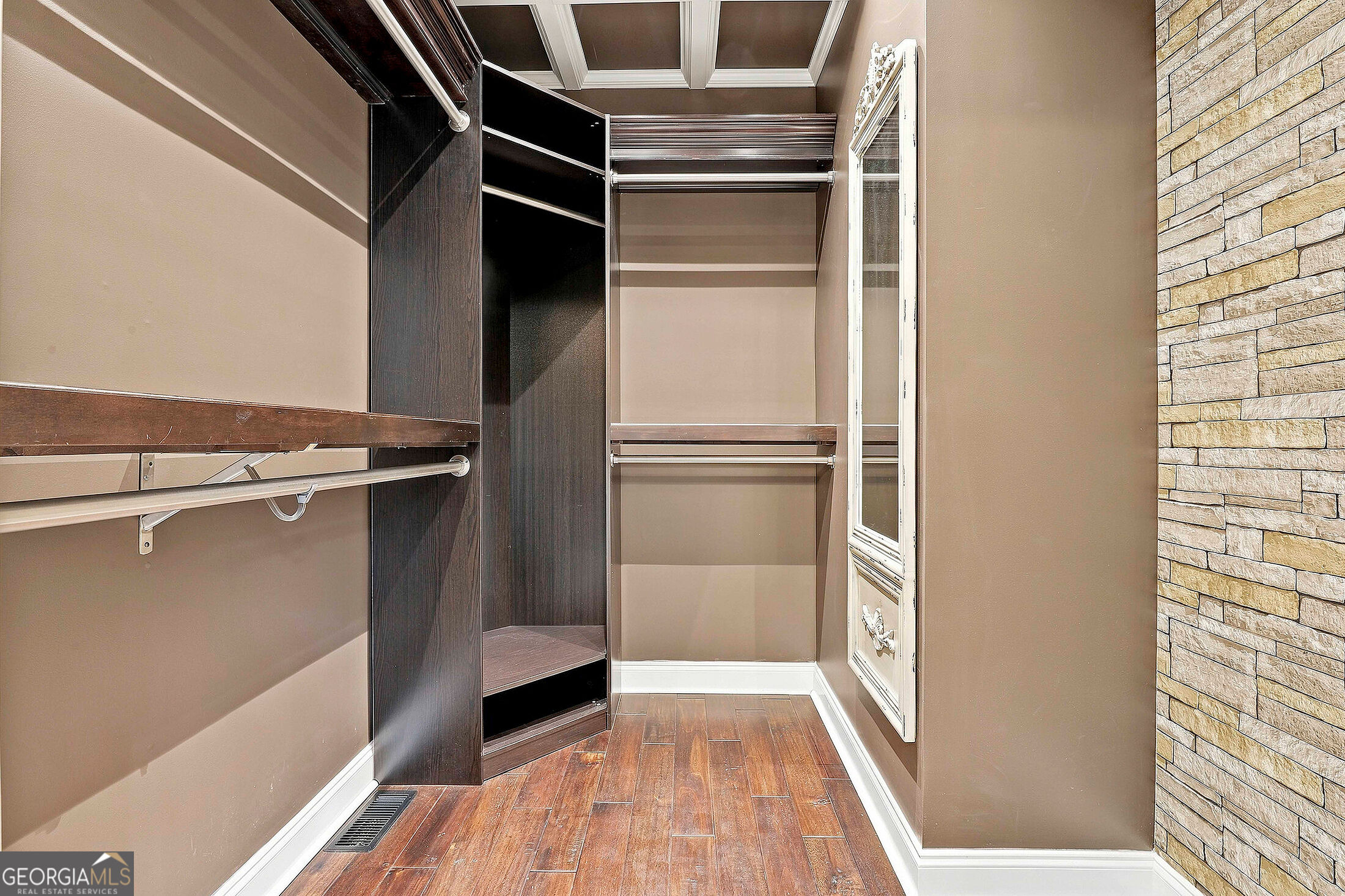 Walk-in closet with dark wood shelving, hanging rods, mirrored cabinet, and a stone accent wall.
