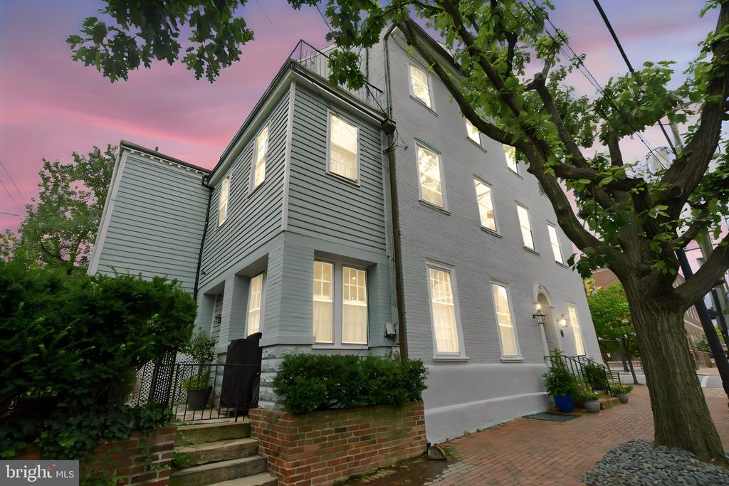 Tall gray house with glowing windows, brick walkway, and a large tree at dusk.