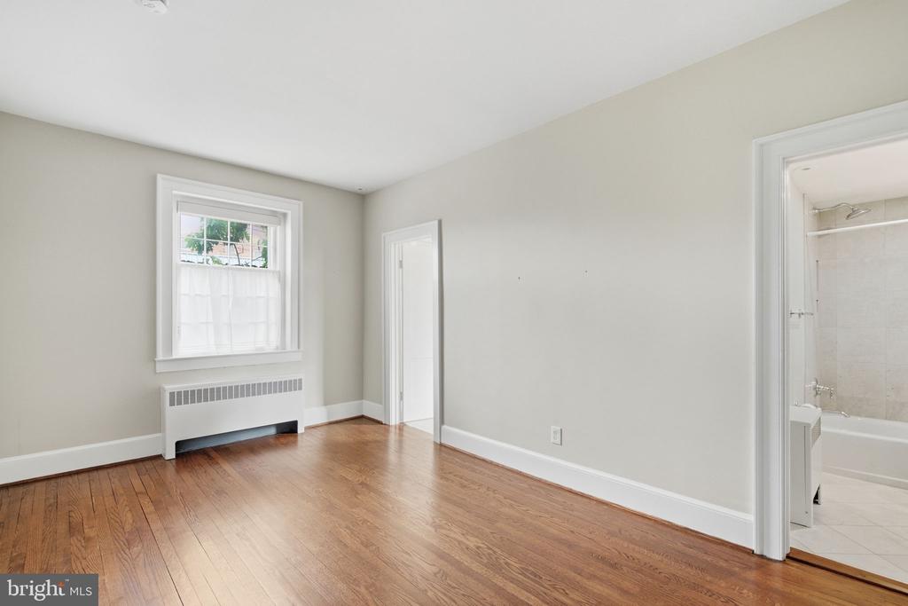 Vacant bedroom with hardwood floors, a window above a radiator, and a bathroom doorway.
