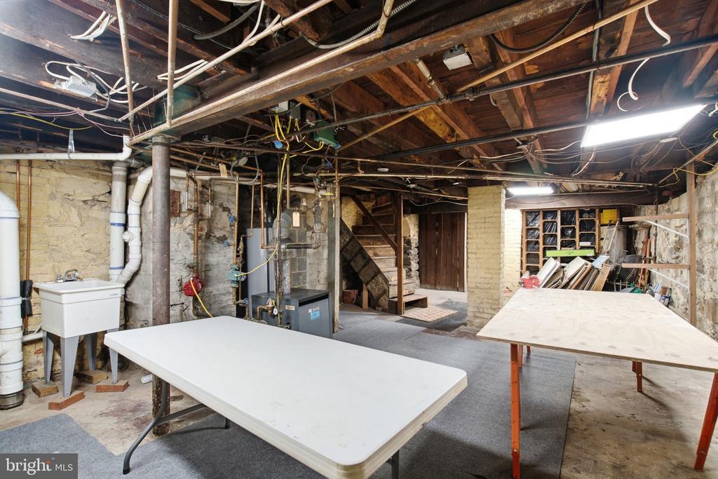 Unfinished basement with exposed beams, stone walls, utility equipment, and large worktables.