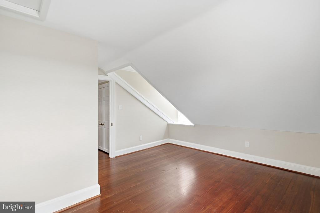 Empty attic room with a sloped ceiling, hardwood floors, and a small triangular window.