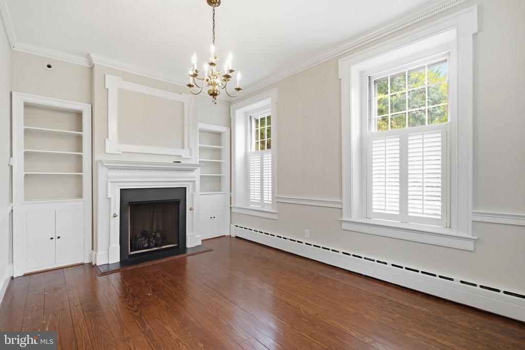 Empty living room with a fireplace, built-in shelves, hardwood floors, and tall shuttered windows.