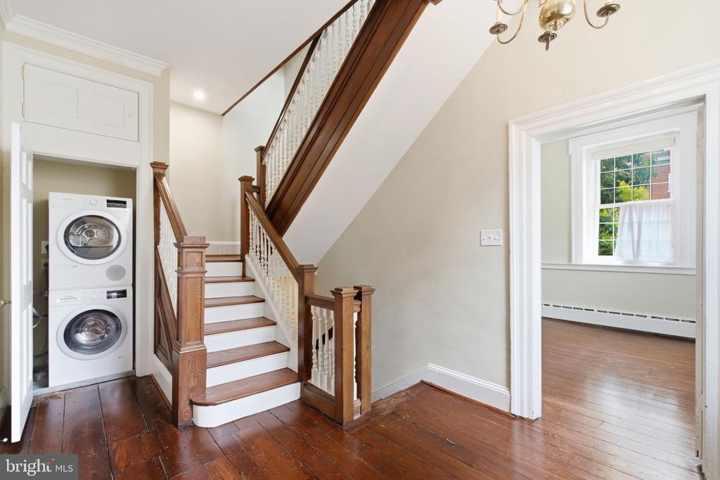 Entry hall with a wood staircase, stacked washer and dryer, and doorway to a bright adjoining room.