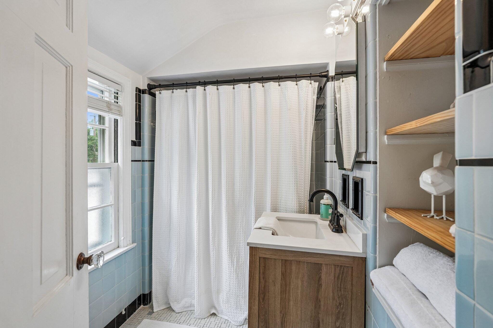 Bathroom with pale blue tile, white shower curtain, wood vanity, and open towel shelves.
