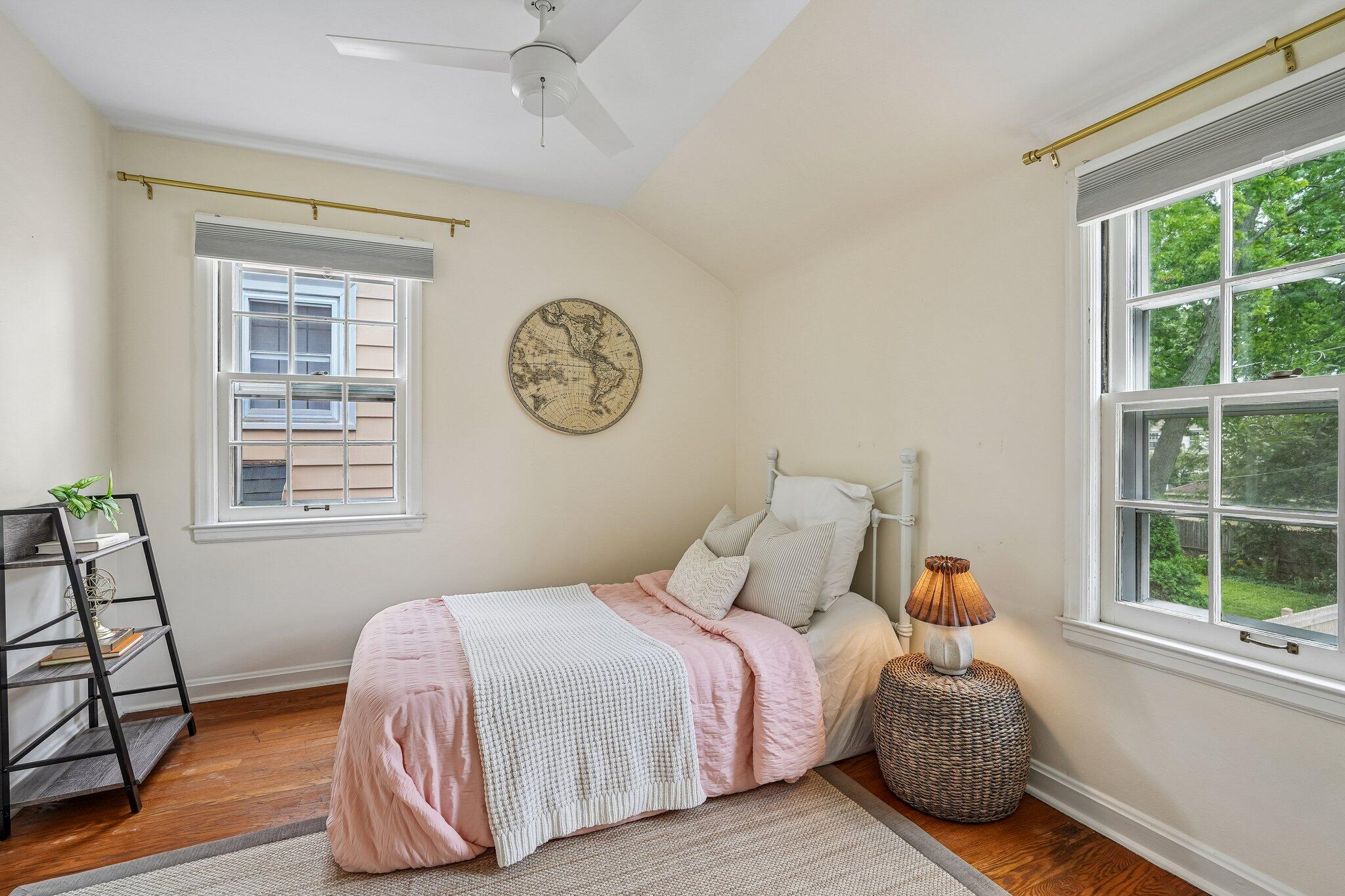 Bedroom with a pink-and-white bed, hardwood floors, and two large windows overlooking greenery.