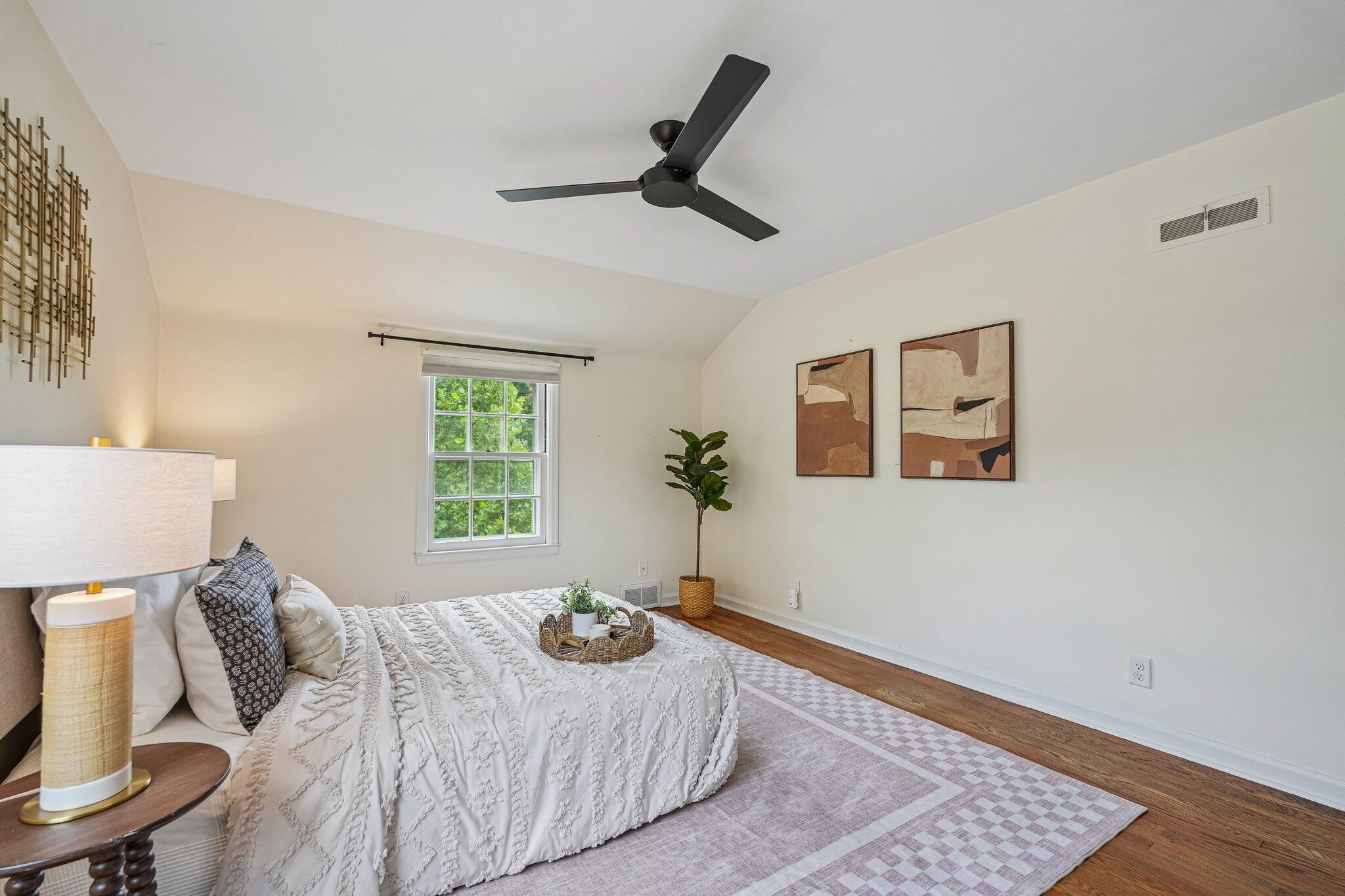 Staged bedroom with a white quilted bed, black ceiling fan, and window overlooking greenery.