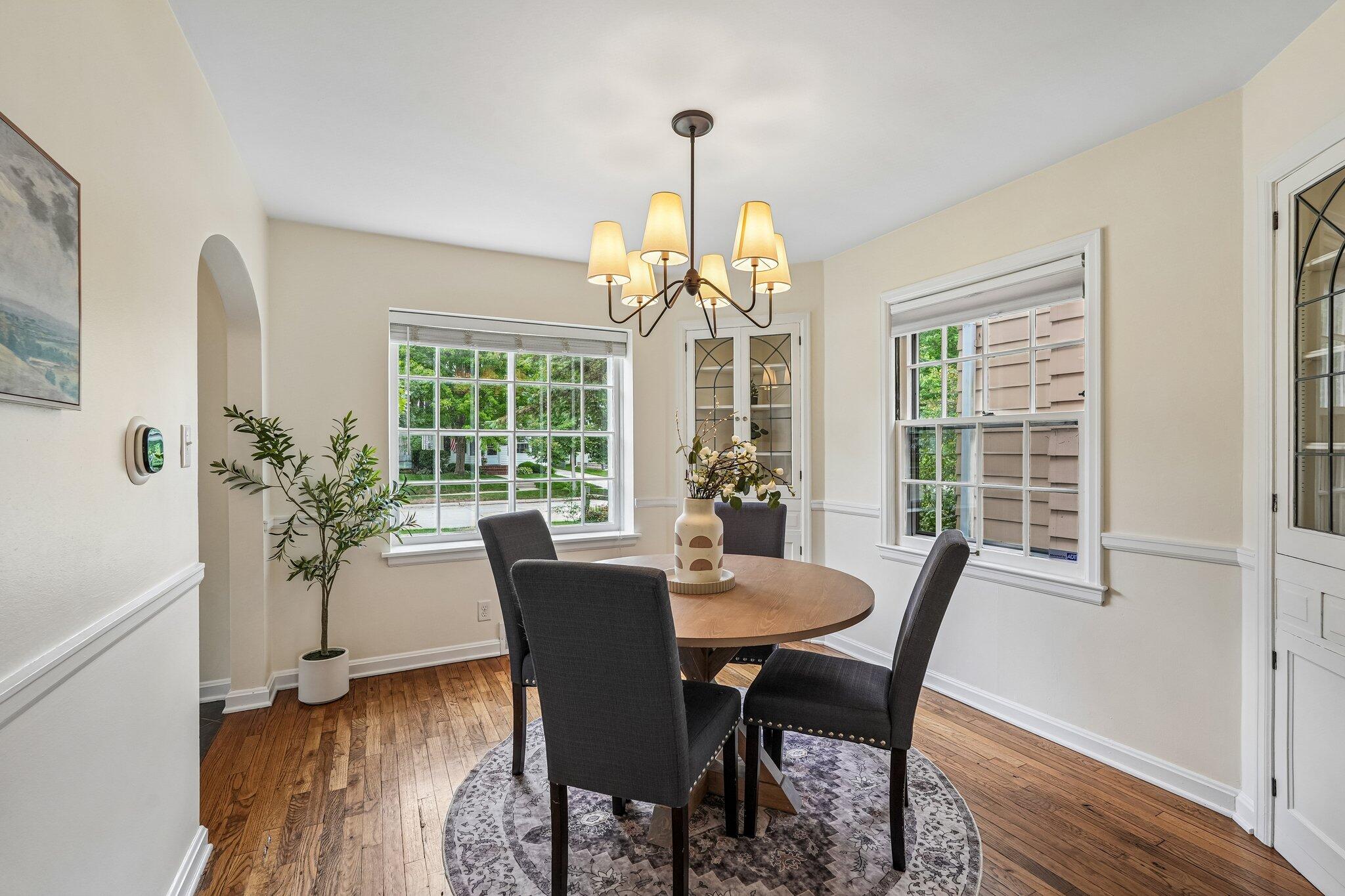 Dining room with a round wood table, dark chairs, and chandelier beneath large windows.