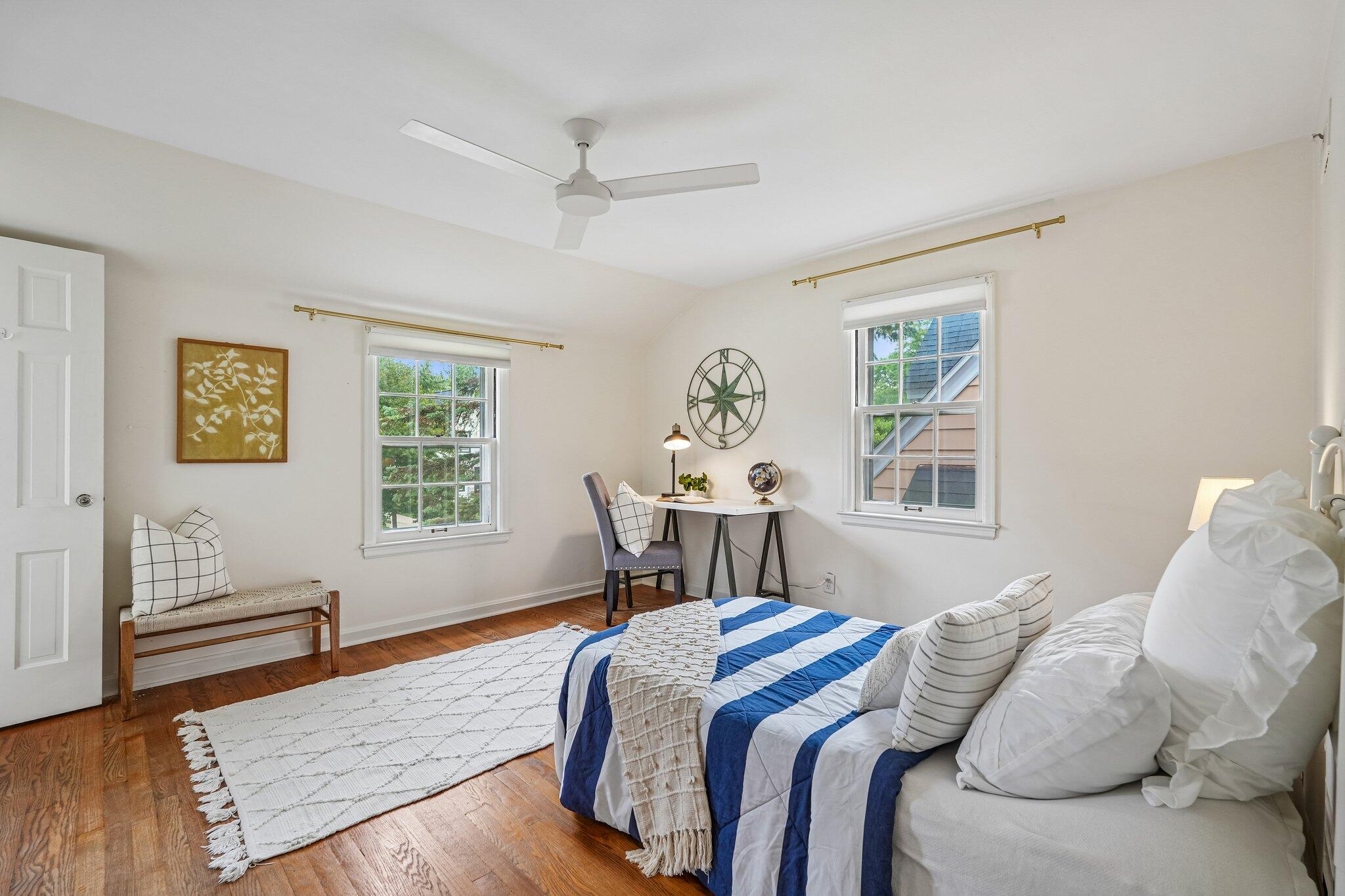 Bedroom with blue-and-white striped bedding, hardwood floors, and a desk between two windows.