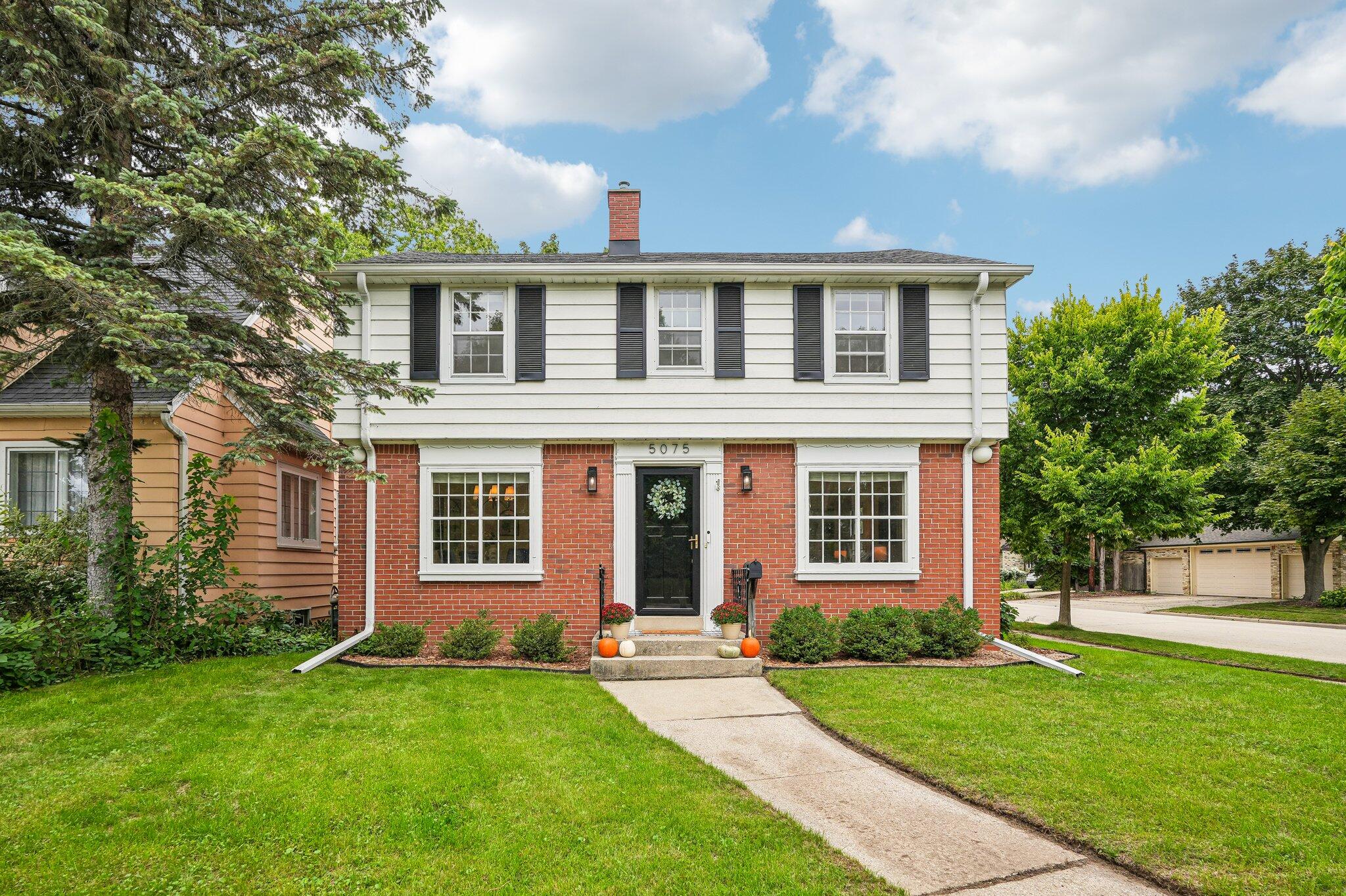 Two-story brick-and-siding house with black shutters, a front walkway, and pumpkins by the entry.