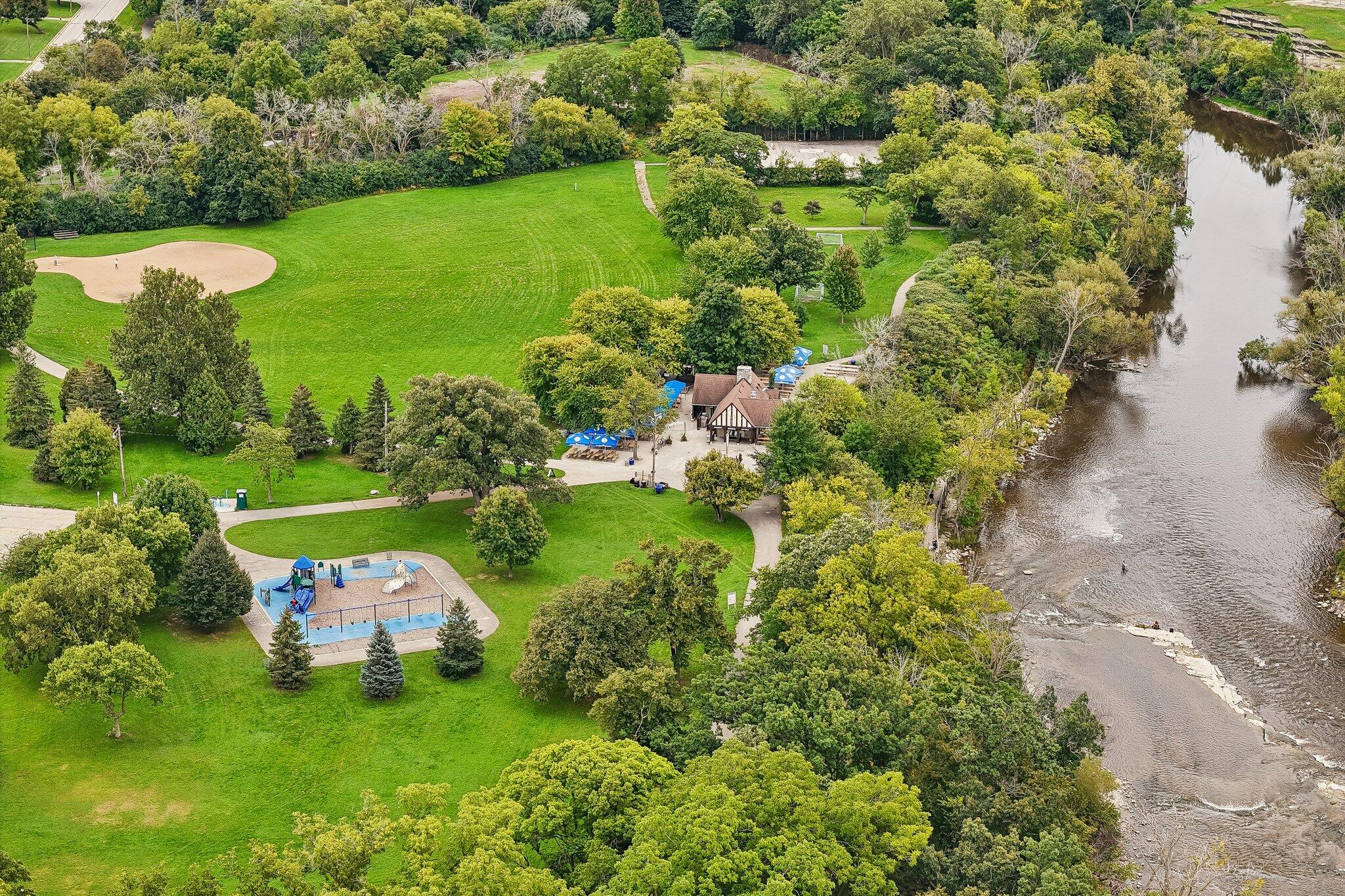 Aerial view of a riverside park with a playground, pavilion, and expansive lawn.