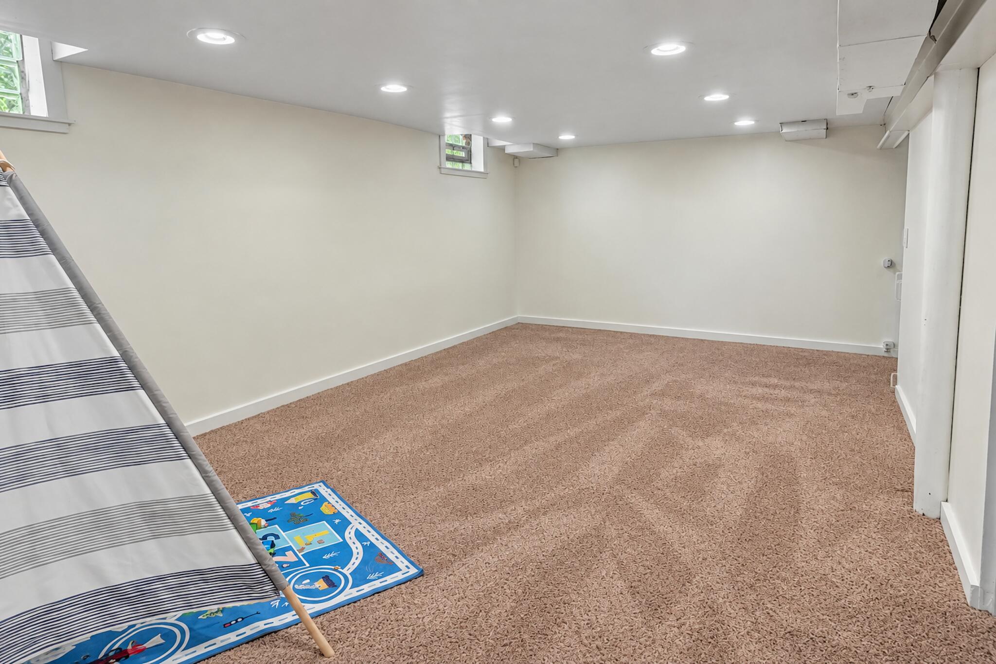 Carpeted basement room with recessed lights, small high windows, and a striped play tent beside a road-pattern mat.