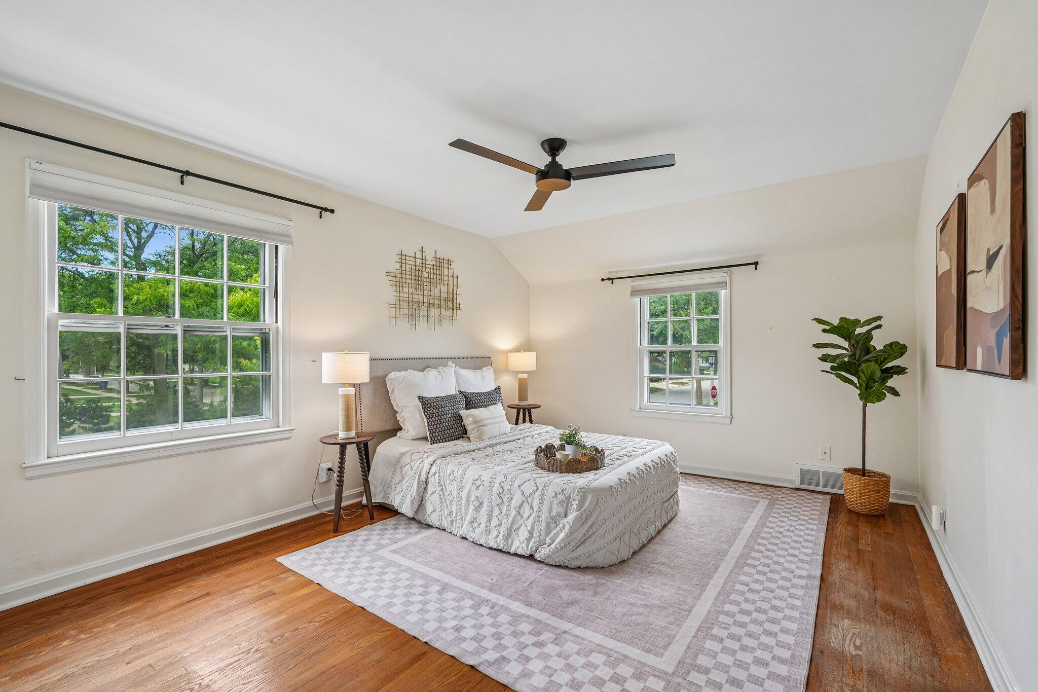 Staged bedroom with a white bed, warm hardwood floors, and large windows overlooking leafy trees.