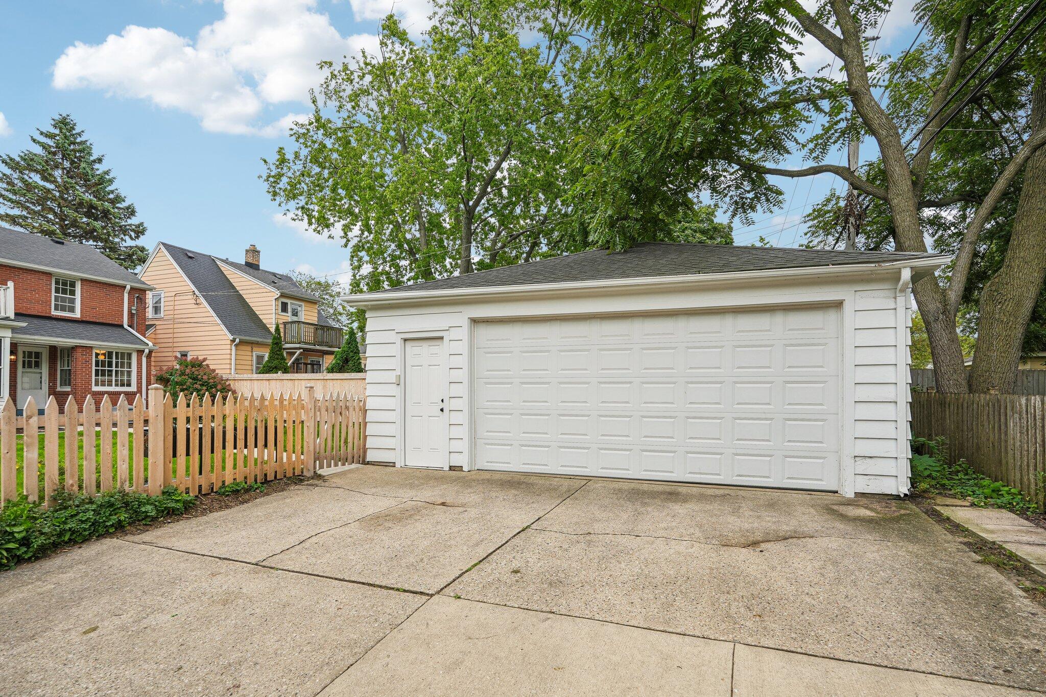 White detached garage with a wide double door, concrete driveway, and picket fence.