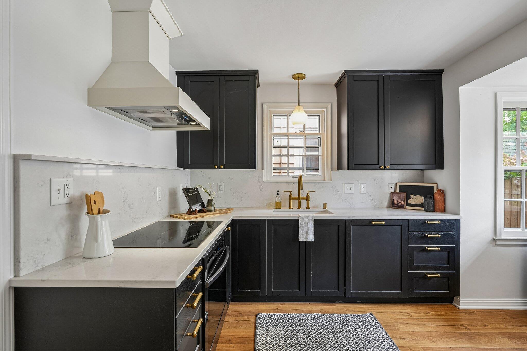 Kitchen with charcoal cabinets, white stone counters, brass fixtures, and a window above the sink.