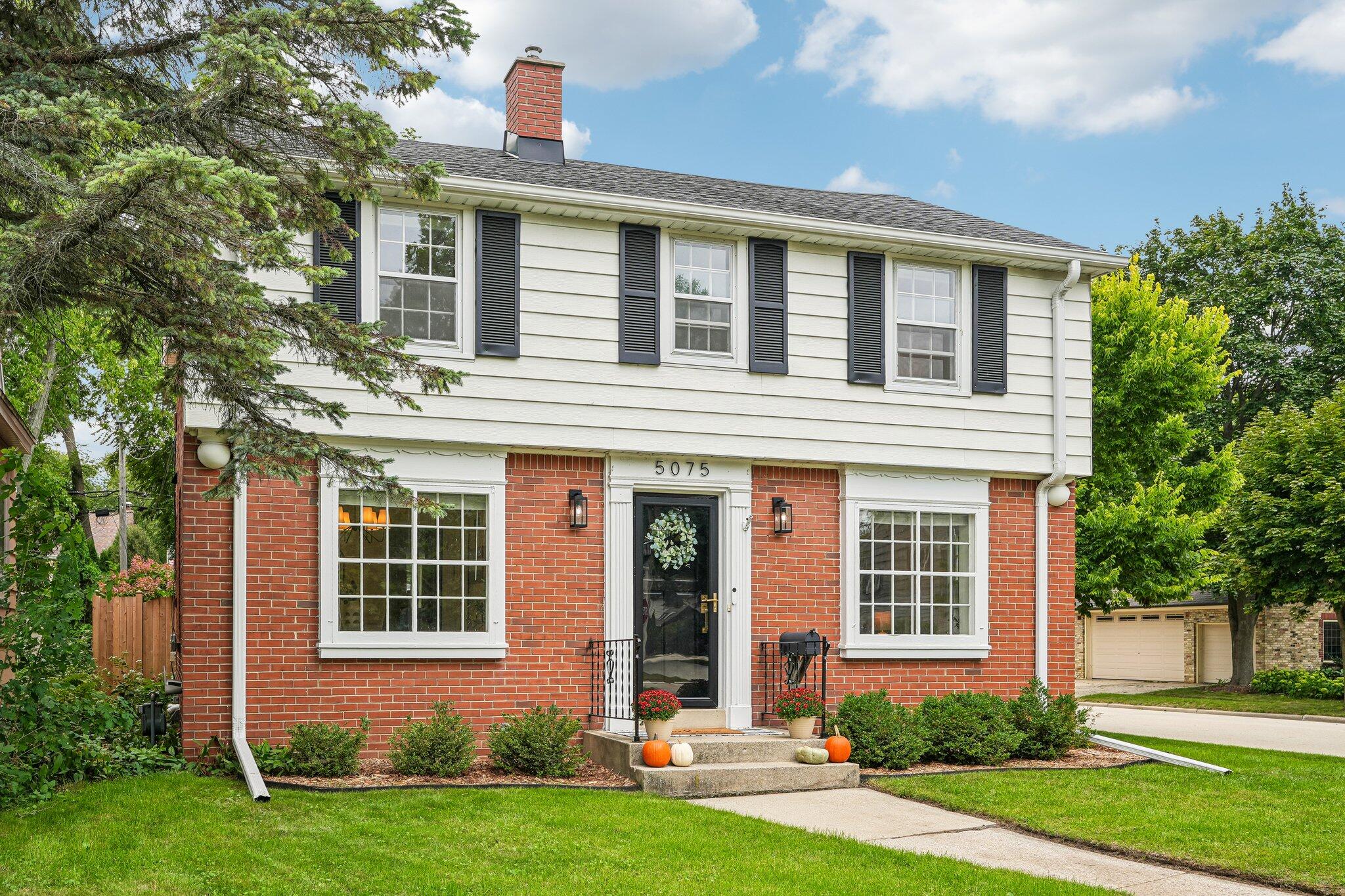 Two-story brick and white-sided home with black shutters, front steps, and pumpkins by the entry.