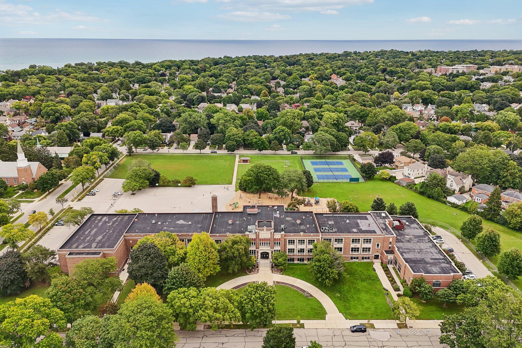 Aerial view of a brick school, tennis courts, playground, and leafy neighborhood beside the water.