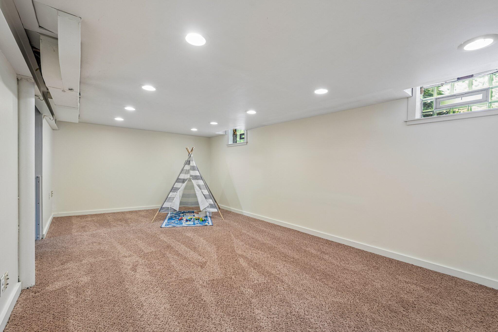 Carpeted lower-level room with a striped teepee and play mat beneath high windows.