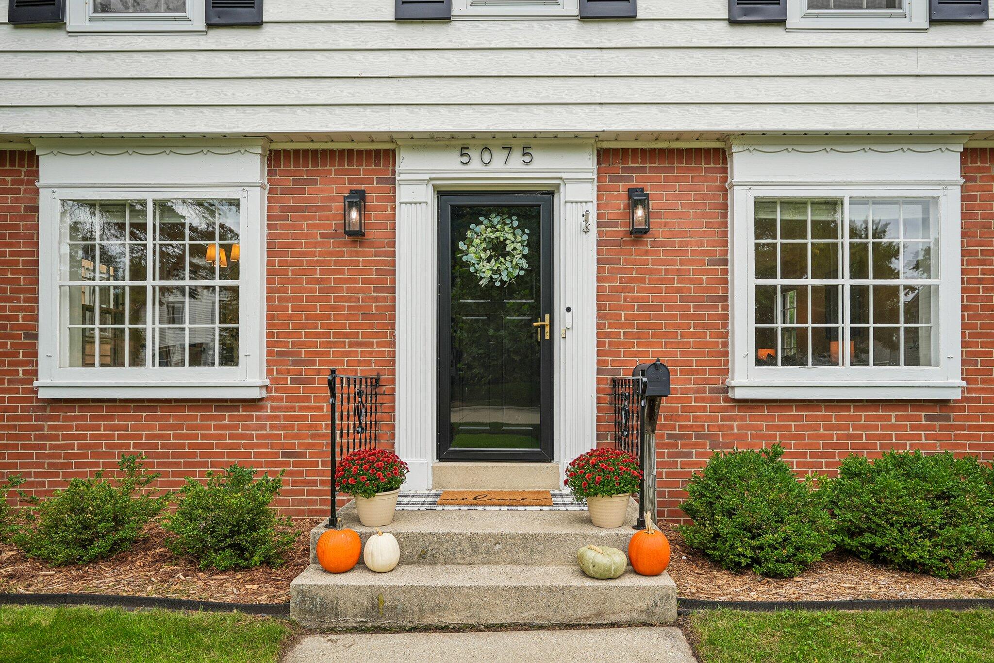 Red-brick home entrance with a black glass door, wreath, flower pots, and pumpkins on the steps.