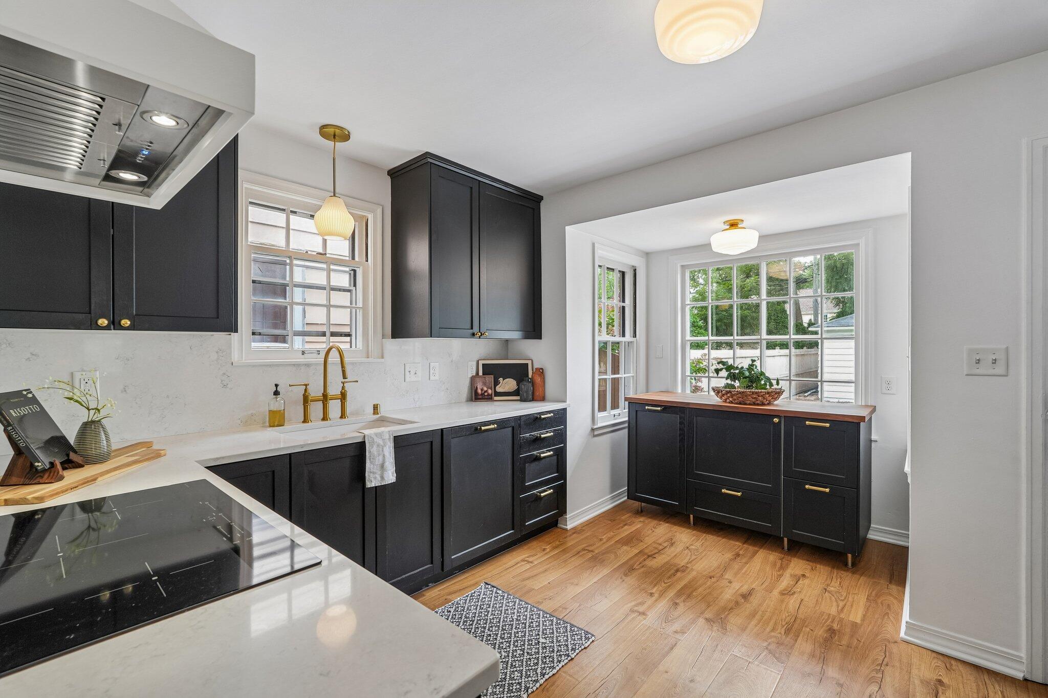 Bright kitchen with charcoal cabinets, white counters, brass fixtures, and a wood-topped cabinet beneath large windows.