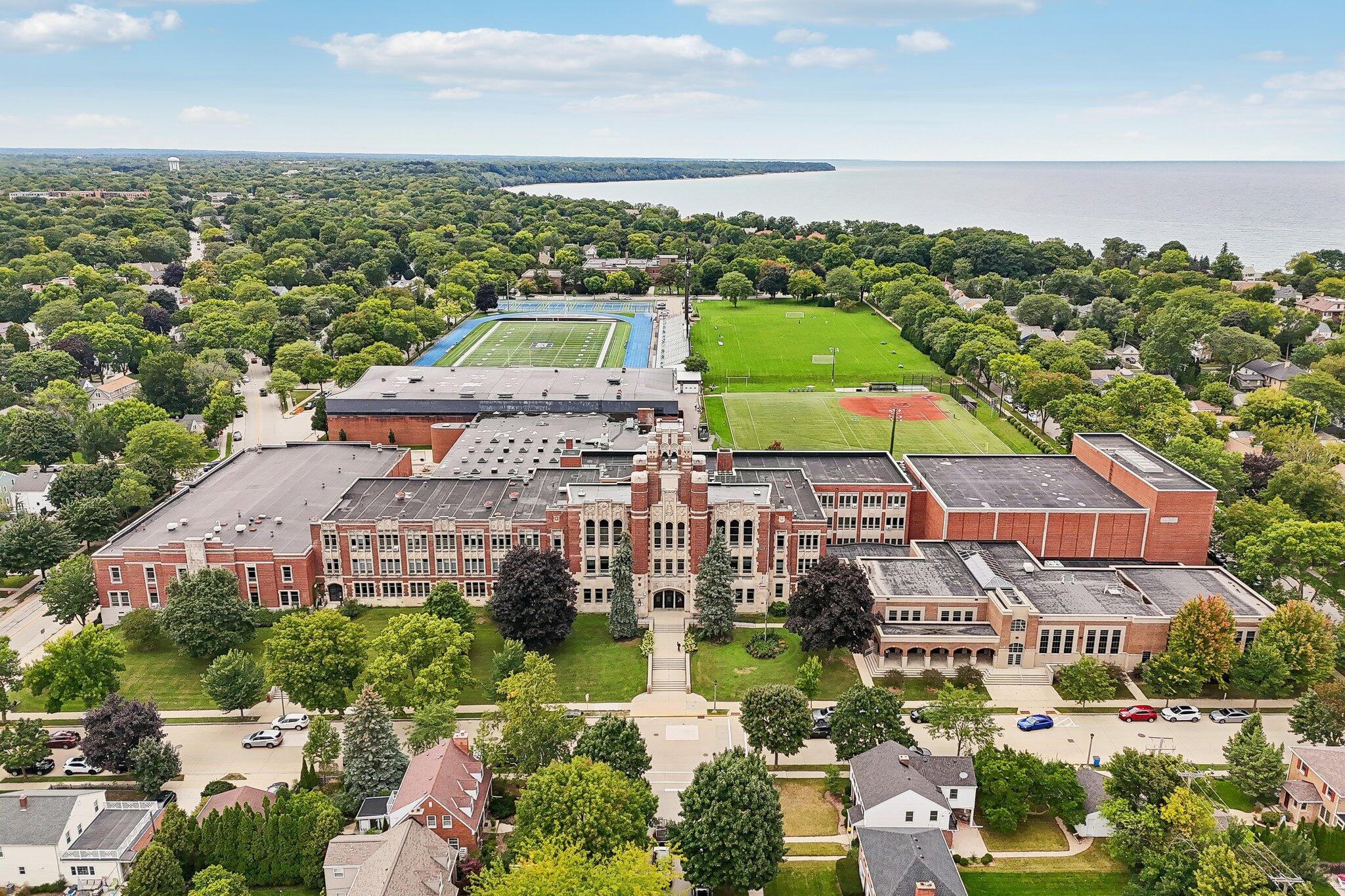 Aerial view of a red-brick school, sports fields, and running track beside a leafy neighborhood and lake.