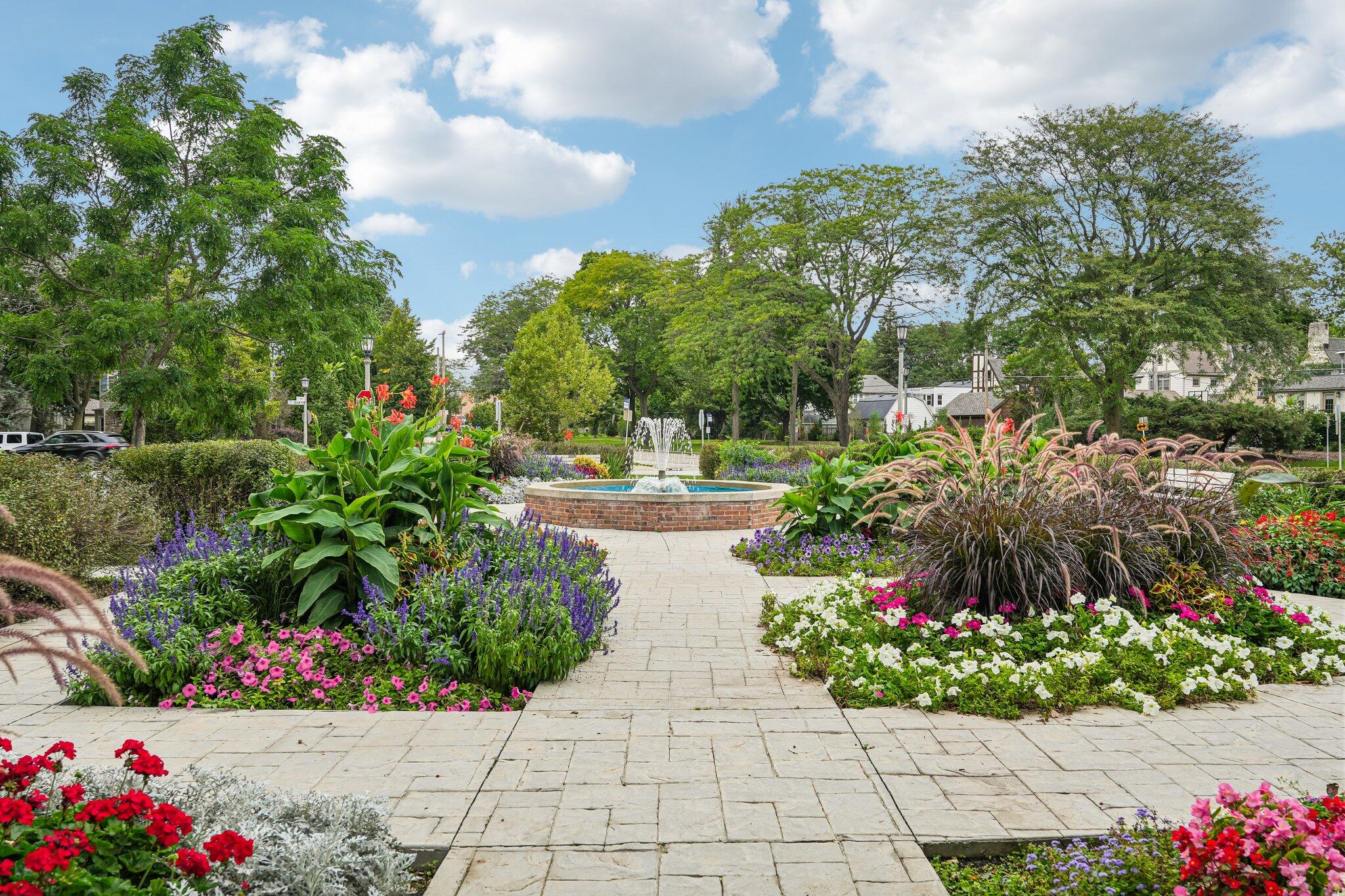Flower gardens surround a brick fountain in a landscaped plaza beneath mature trees.