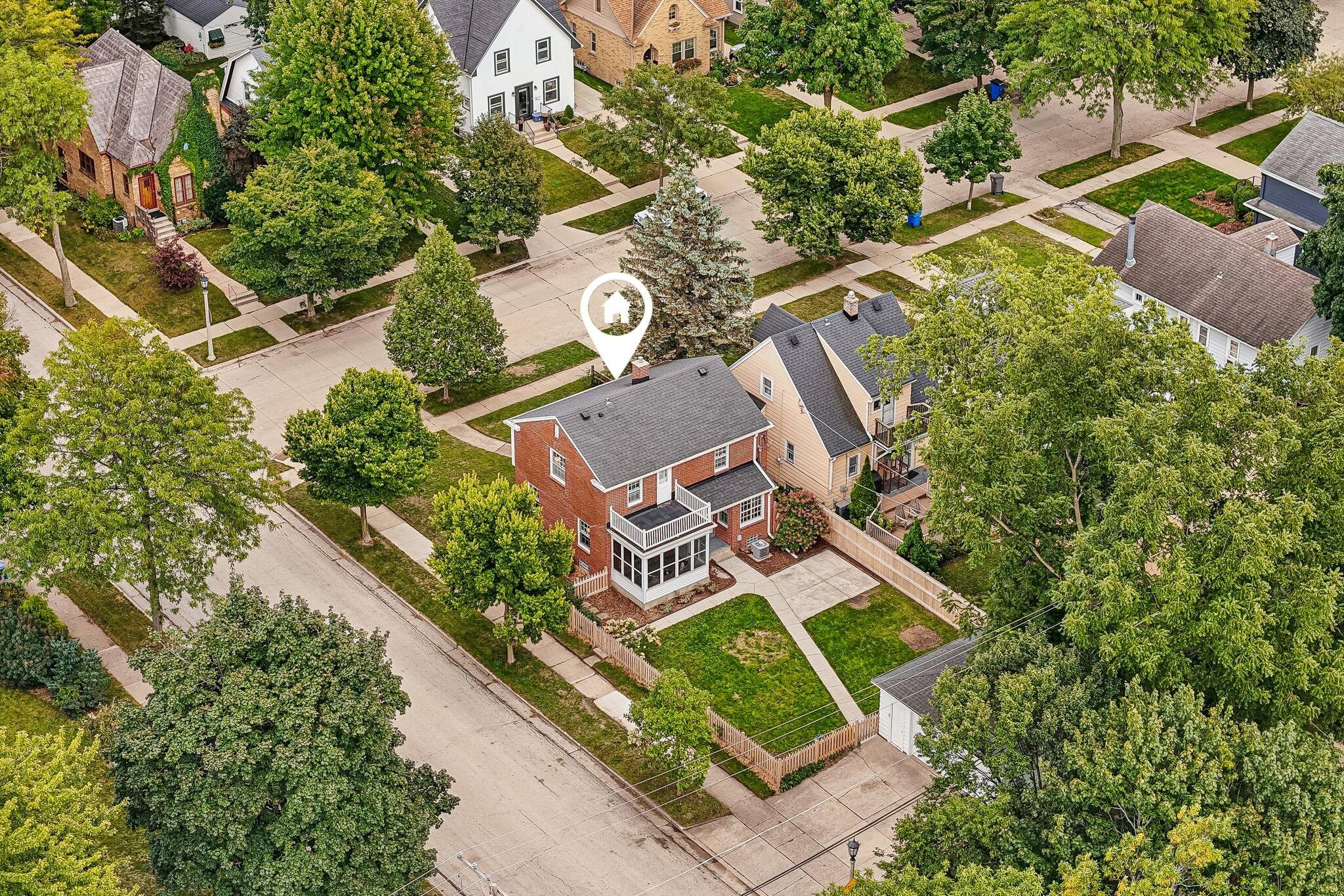 Aerial view of a red-brick house with an enclosed porch and fenced lawn on a tree-lined street.