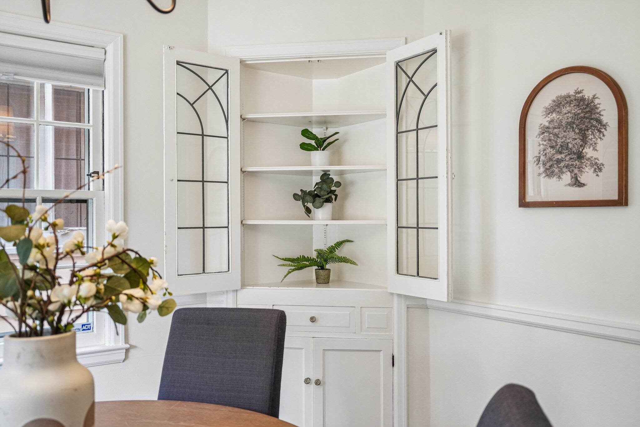Dining area with an open white corner cabinet, glass-paneled doors, and plants on the shelves.