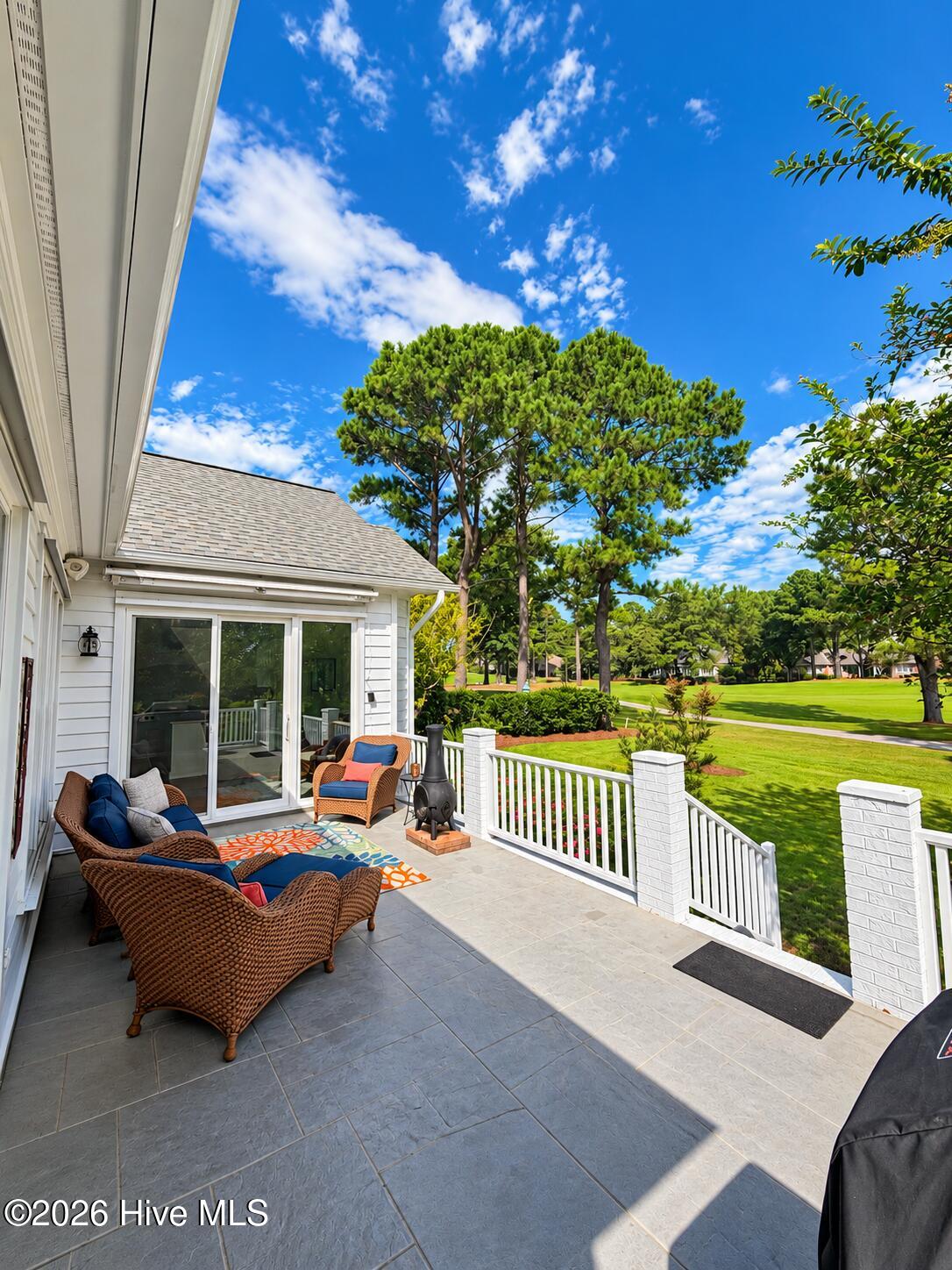 Tiled patio with wicker seating and white railing overlooking a landscaped lawn and pine trees.