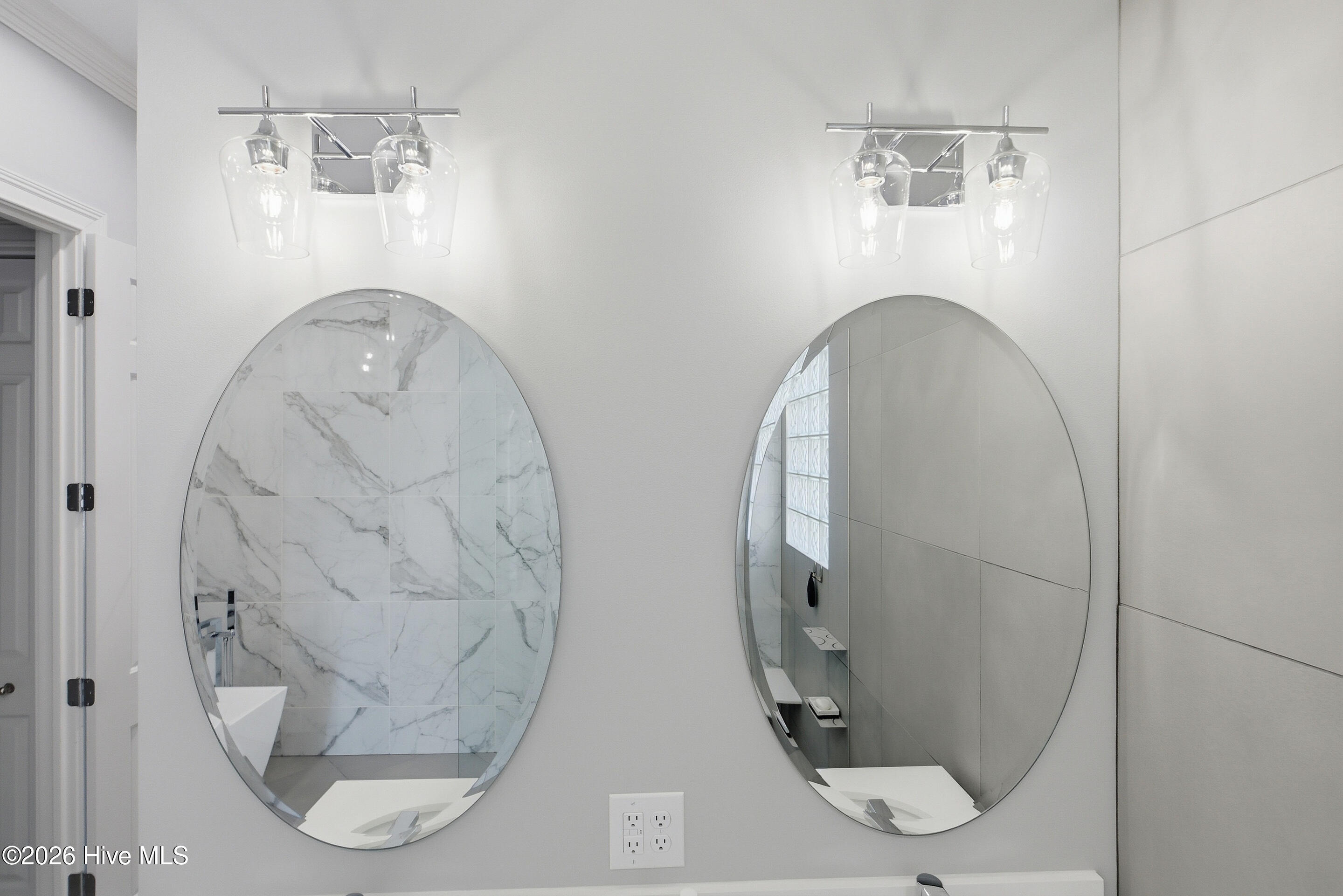 Bathroom vanity with twin oval mirrors, chrome lights, and marble-look tile reflected in the mirrors.