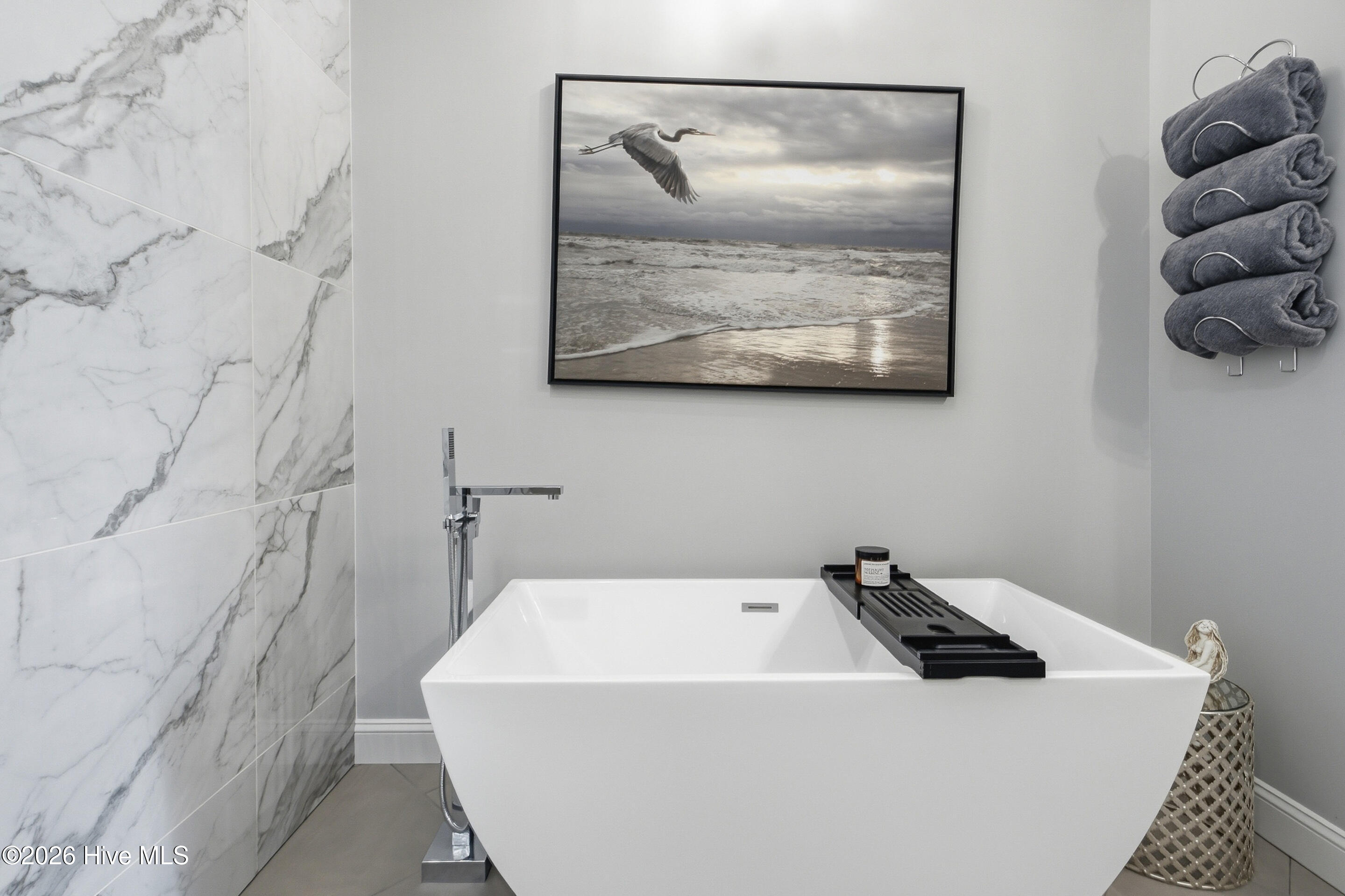 Modern bathroom with a freestanding white tub, marble-look tile, and framed heron beach artwork.